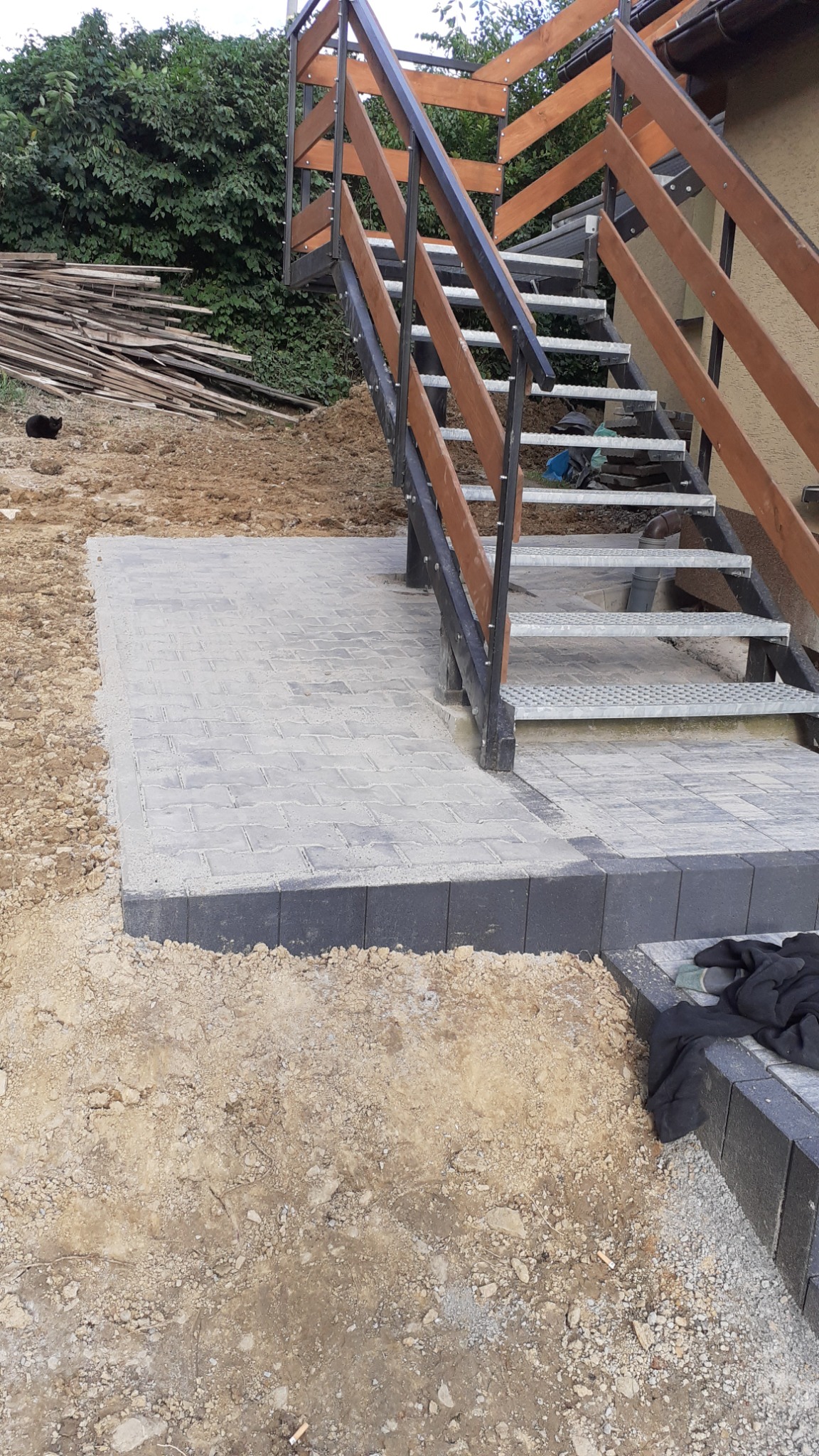 Świeżo ułożony chodnik z szarej kostki brukowej przy metalowych schodach z drewnianą balustradą, widoczne pozostałości piasku i ziemi po pracach, w tle sterta desek i zielona roślinność.
