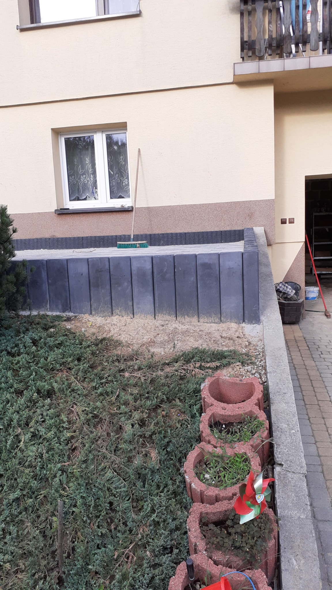 Świeżo wykończony murek oporowy z betonowych elementów, zabezpieczający skarpę przy budynku, z widoczną szczotką do zamiatania na górze i rzędem czerwonych doniczek w kształcie serca wzdłuż...