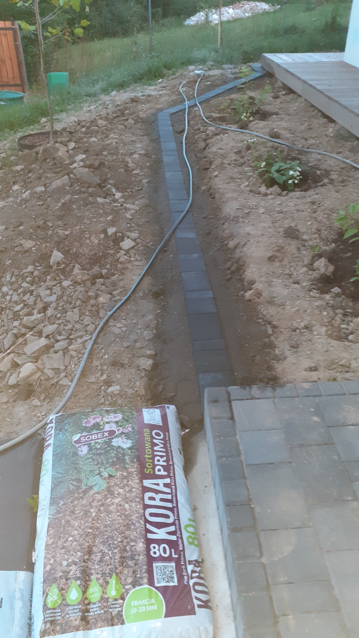 Wykopany rów w ziemi z ułożonymi obrzeżami z kostki brukowej, widoczny kabel i worek kory sosnowej 80L w tle, przygotowanie do dalszych prac w ogrodzie.