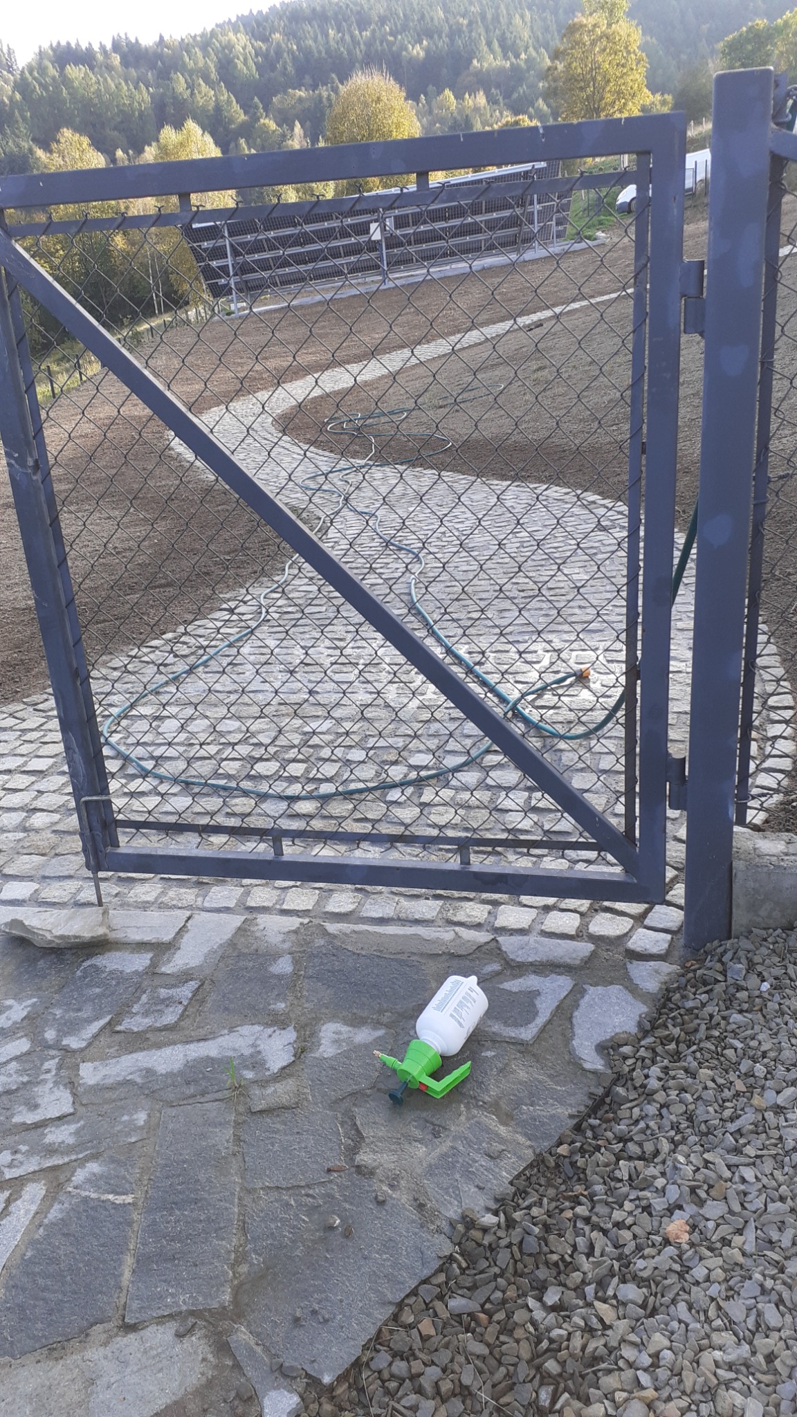 Widok przez metalową furtkę na świeżo wyrównany teren pod trawnik, z widocznym systemem nawadniania i butelką opryskiwacza leżącą na kamiennej ścieżce.