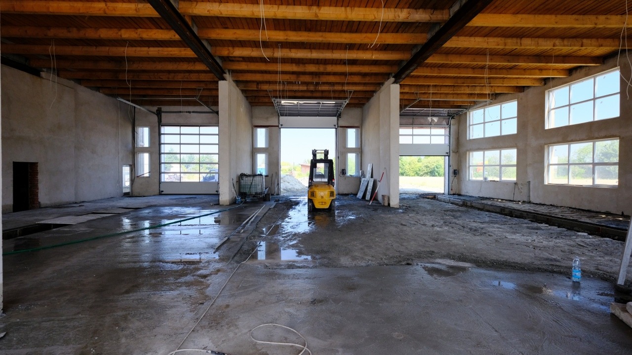 Wnętrze hali w trakcie prac wykończeniowych, widoczny surowy beton, drewniany strop, okna z białymi ramami, otwarte bramy garażowe oraz żółty wózek widłowy w oddali.