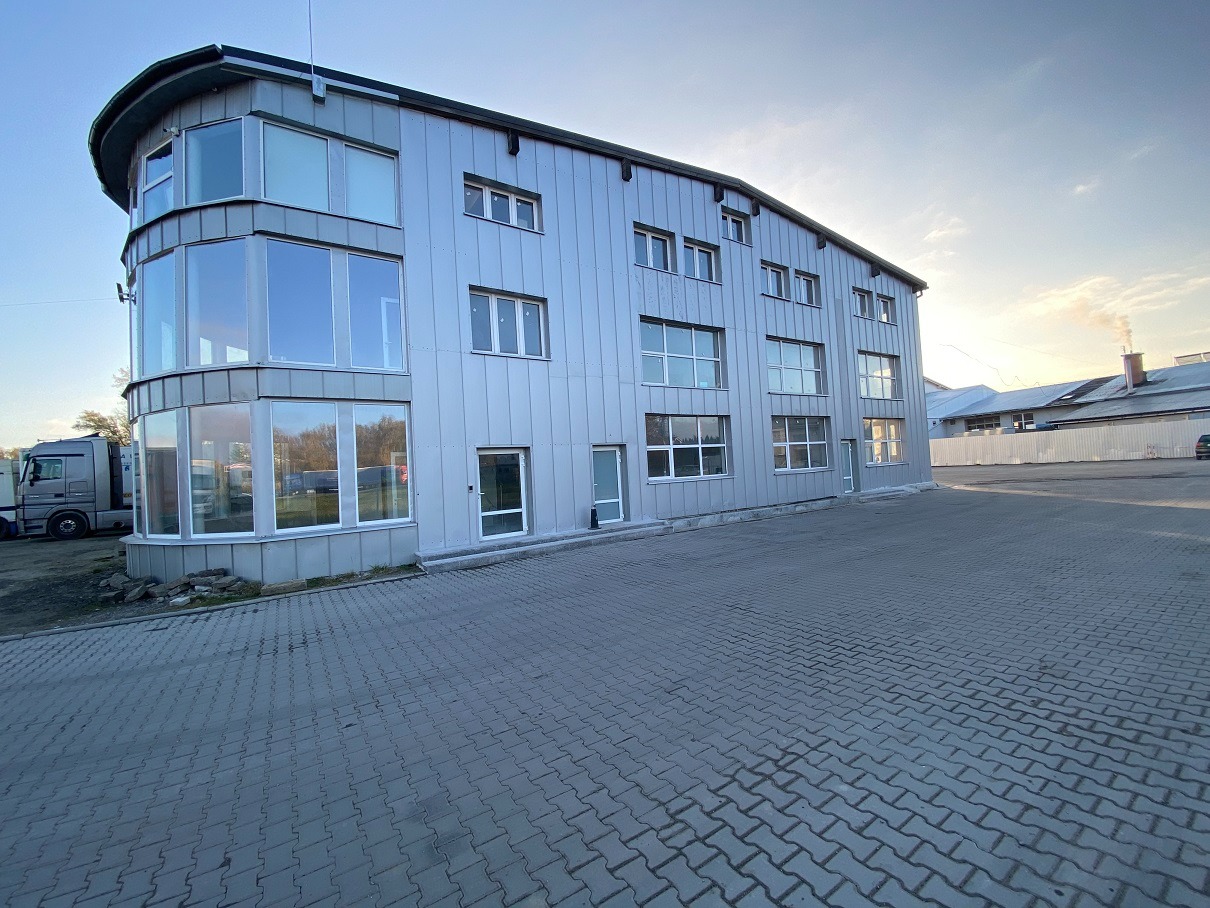 Trzykondygnacyjny budynek przemysłowy z elewacją z blachy trapezowej i panoramicznym narożnikiem z dużymi oknami, widok z zewnątrz na kostkę brukową i zaparkowany samochód ciężarowy.