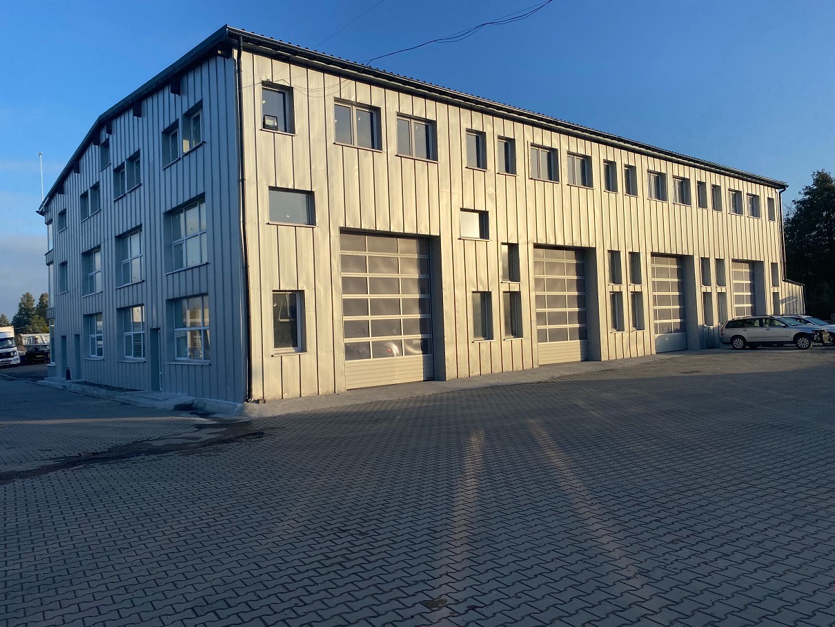 Duży budynek przemysłowy z szarej blachy trapezowej, widoczne okna i segmentowe bramy garażowe, zaparkowane samochody na zewnątrz.