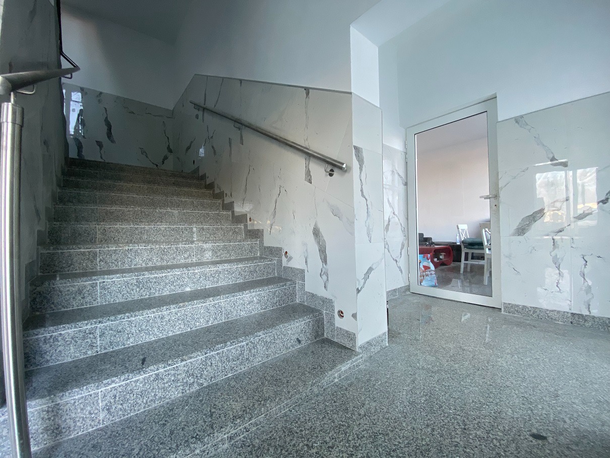 Wykończona klatka schodowa z szarymi granitowymi schodami i białymi marmurkowymi ścianami, widok na białe drzwi z widocznym wnętrzem mieszkania.