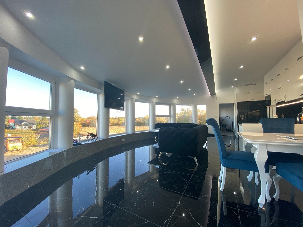 Nowoczesne wnętrze z czarną marmurową podłogą, panoramicznymi oknami z widokiem na krajobraz, ciemnoniebieską sofą chesterfield i białym stołem jadalnym z krzesłami w podobnym kolorze. Widoczny...