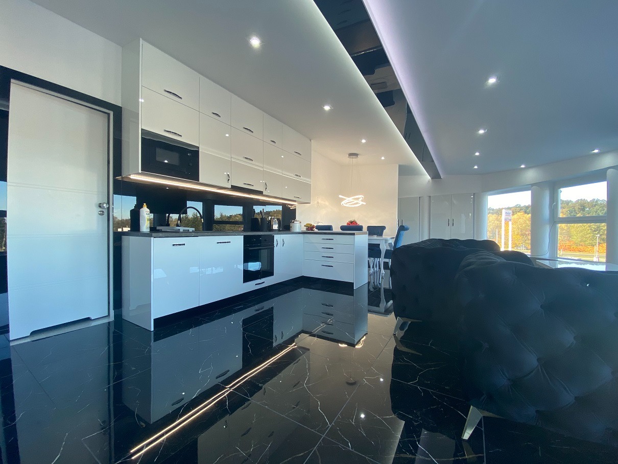 Nowoczesna kuchnia z białymi, lakierowanymi szafkami, czarnym blatem i podświetleniem LED, odbijająca się w czarnej, marmurowej podłodze, widok na salon z ciemną sofą i okrągłym stołem jadalnym.