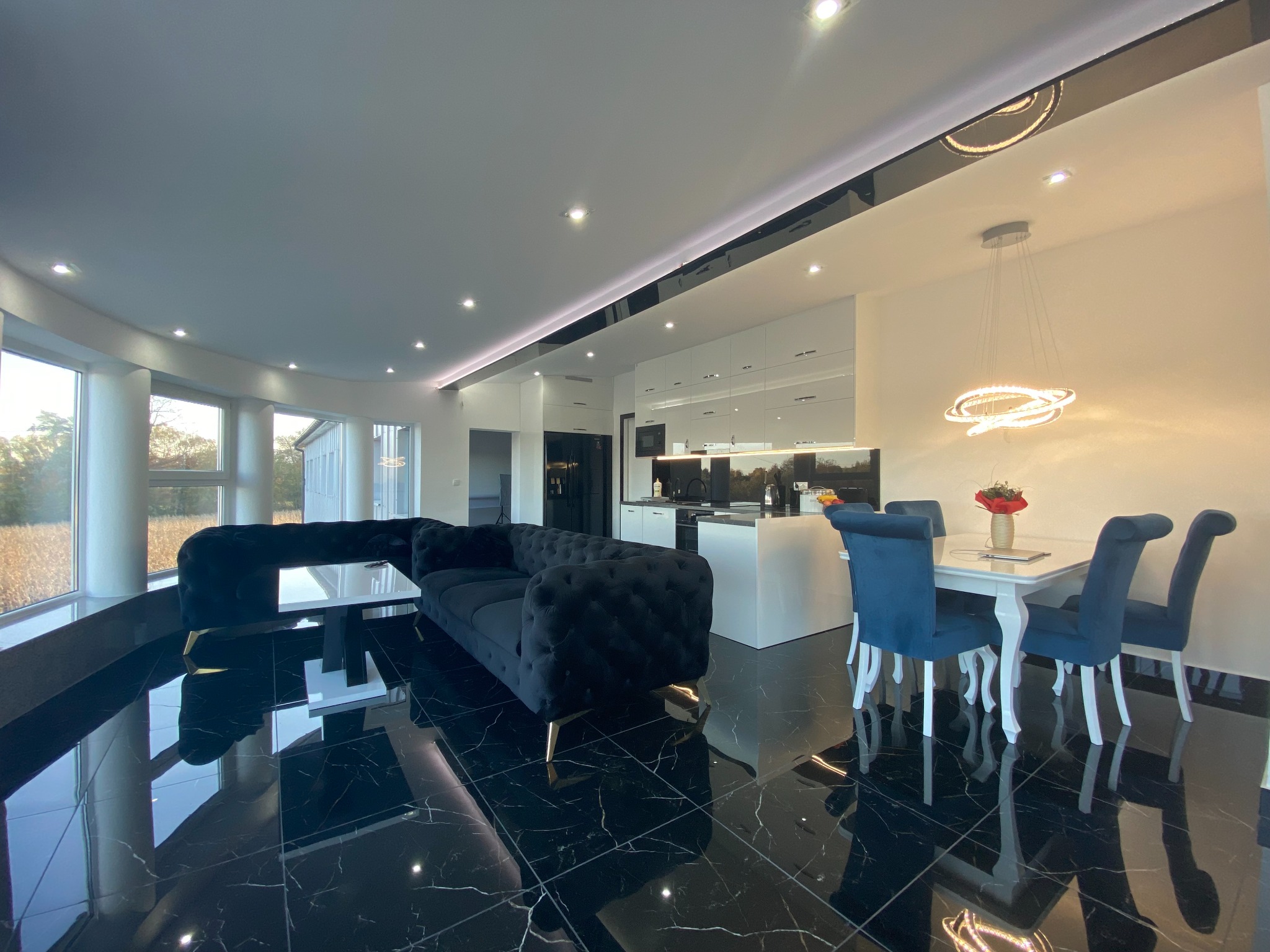 Eleganckie wnętrze domu z czarną, błyszczącą podłogą z płytek, pluszową sofą na złotych nóżkach, białą kuchnią z wyspą i stołem z niebieskimi krzesłami, oświetlone nowoczesnymi lampami.