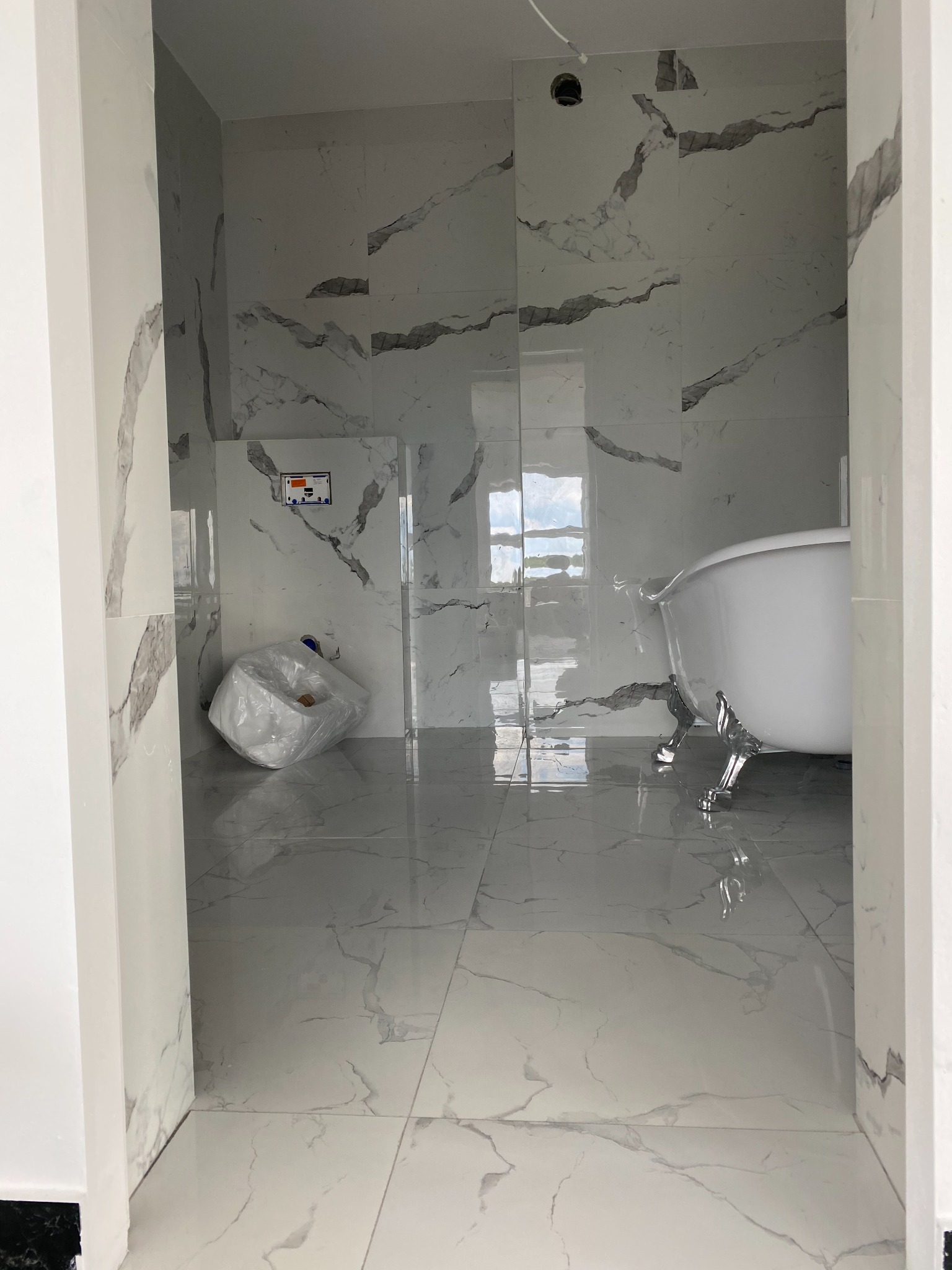 Nowoczesna łazienka w trakcie remontu z białą wanną na lwich nóżkach, marmurowe płytki na ścianach i podłodze, oraz zabezpieczona folią toaleta w rogu.