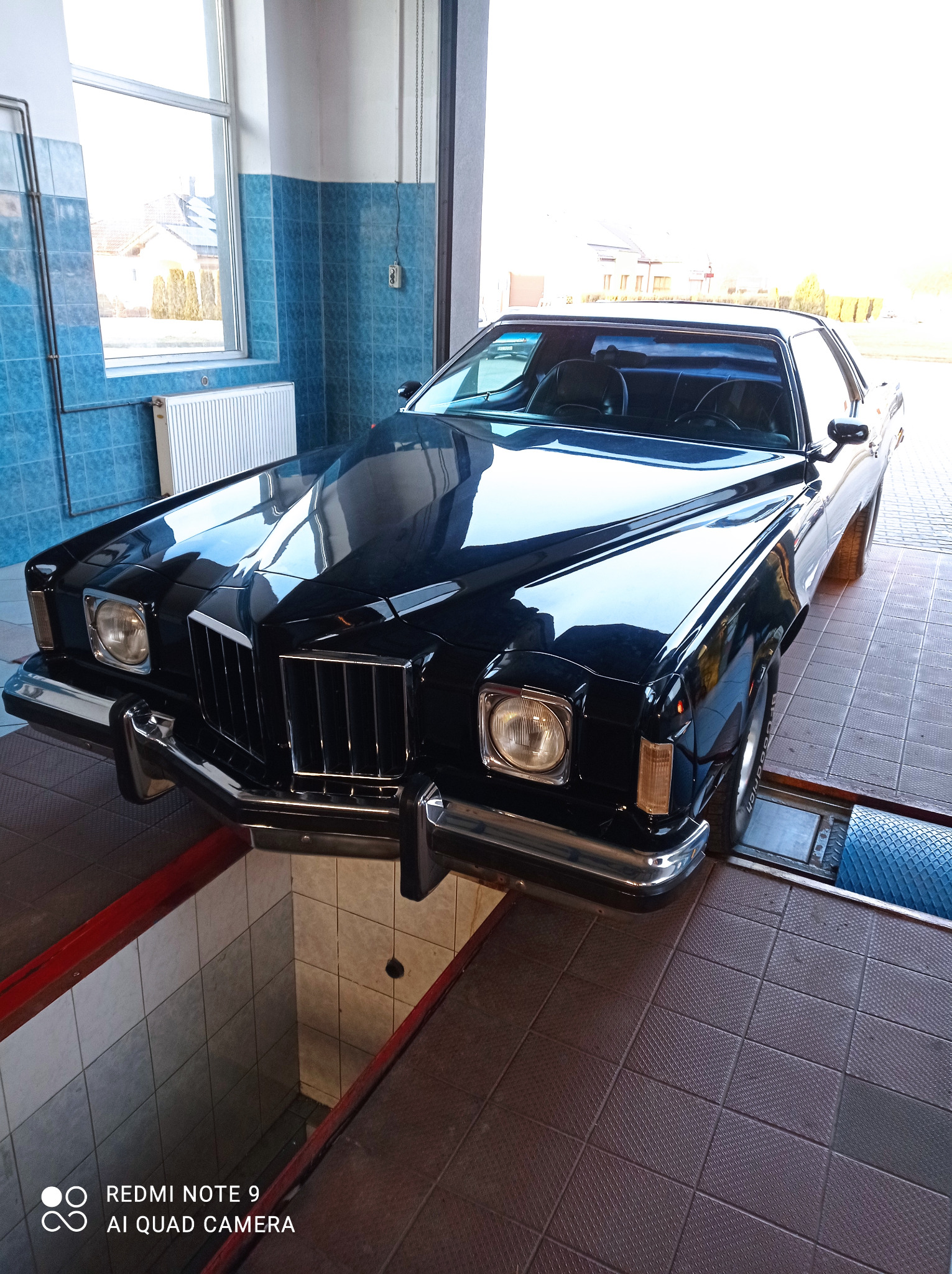 Czarny, klasyczny samochód marki Pontiac Grand Prix z lat 70. na podnośniku w warsztacie samochodowym z niebieskimi kafelkami na ścianach.