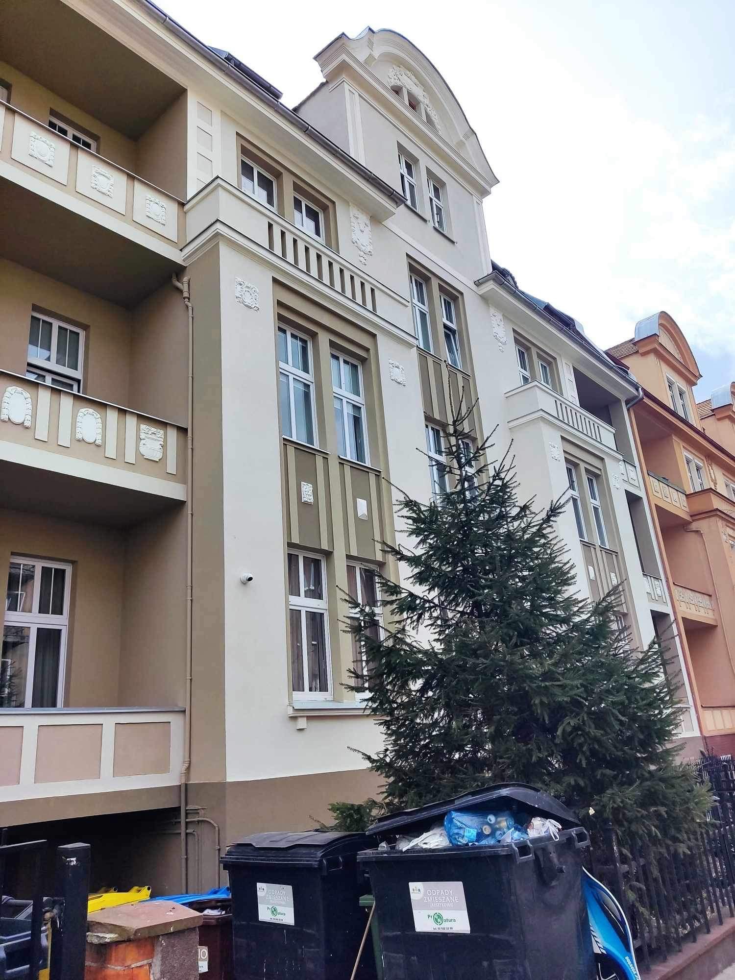 Odnowiona elewacja kamienicy z elementami dekoracyjnymi i balkonami, widoczna z poziomu ulicy, z koszami na śmieci i iglakiem na pierwszym planie.