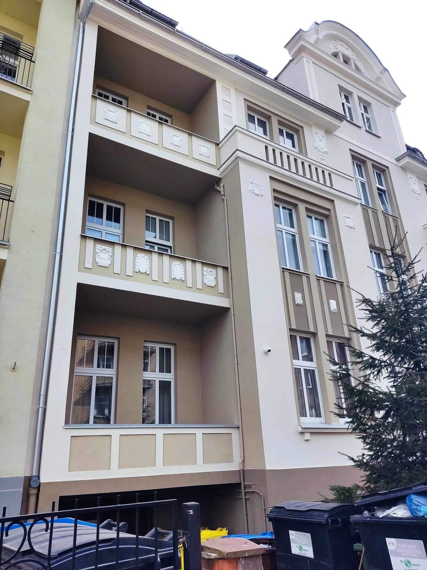 Odnowiona elewacja kamienicy z balkonami ozdobionymi płaskorzeźbami, widok z dołu na fragment budynku z elementami dekoracyjnymi i oknami, z widocznymi śmietnikami w dolnej części kadru.