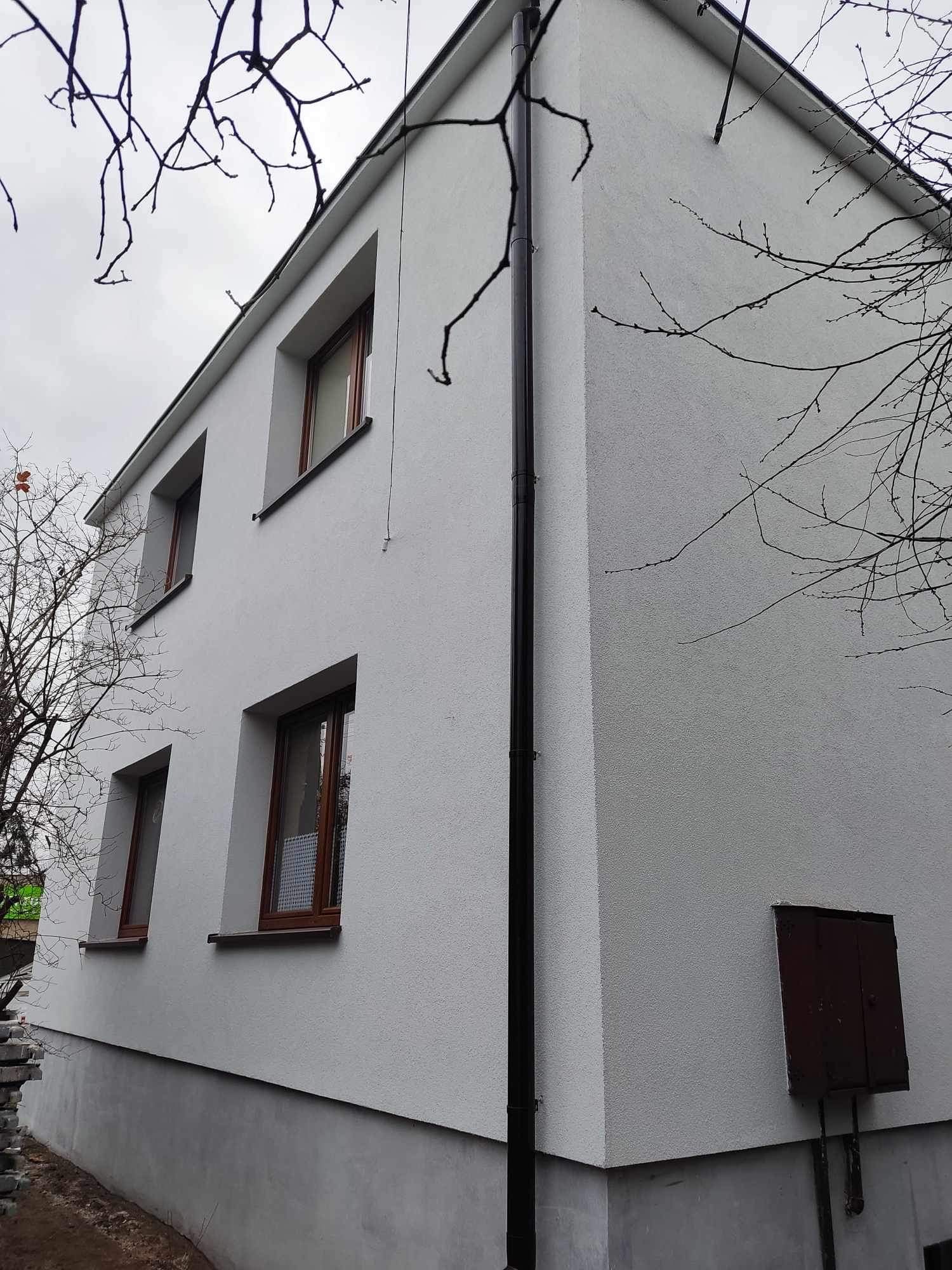 Dwupiętrowy dom z nową, jasnoszarą elewacją o fakturze baranka, brązowe okna i rynna na rogu budynku, widoczne nagie gałęzie drzew