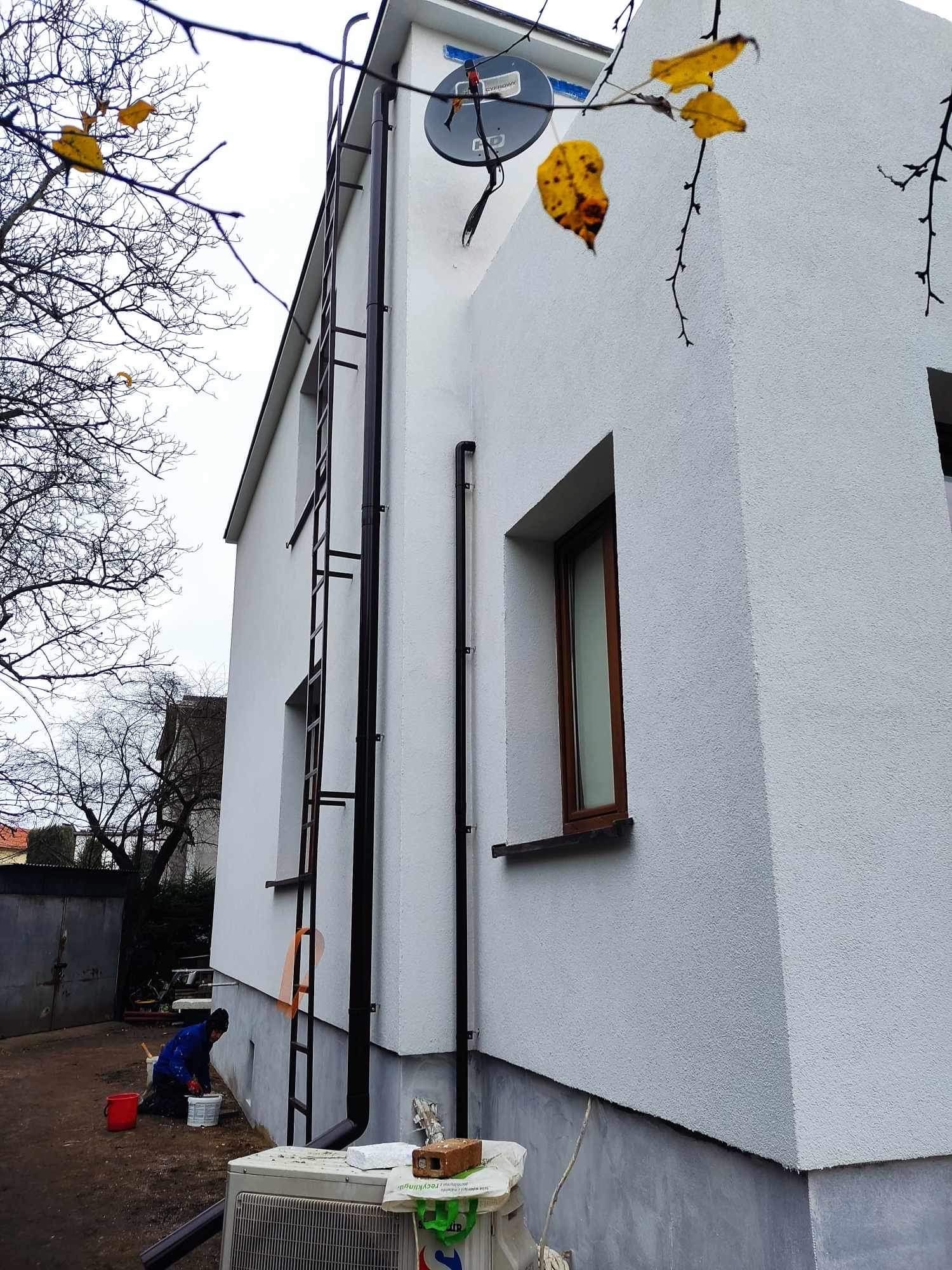 Boczny widok domu z nową białą elewacją o fakturze baranka, z zamontowaną drabinką, rynnami i anteną satelitarną. Pracownik w niebieskim kombinezonie przygotowuje się do prac wykończeniowych przy...