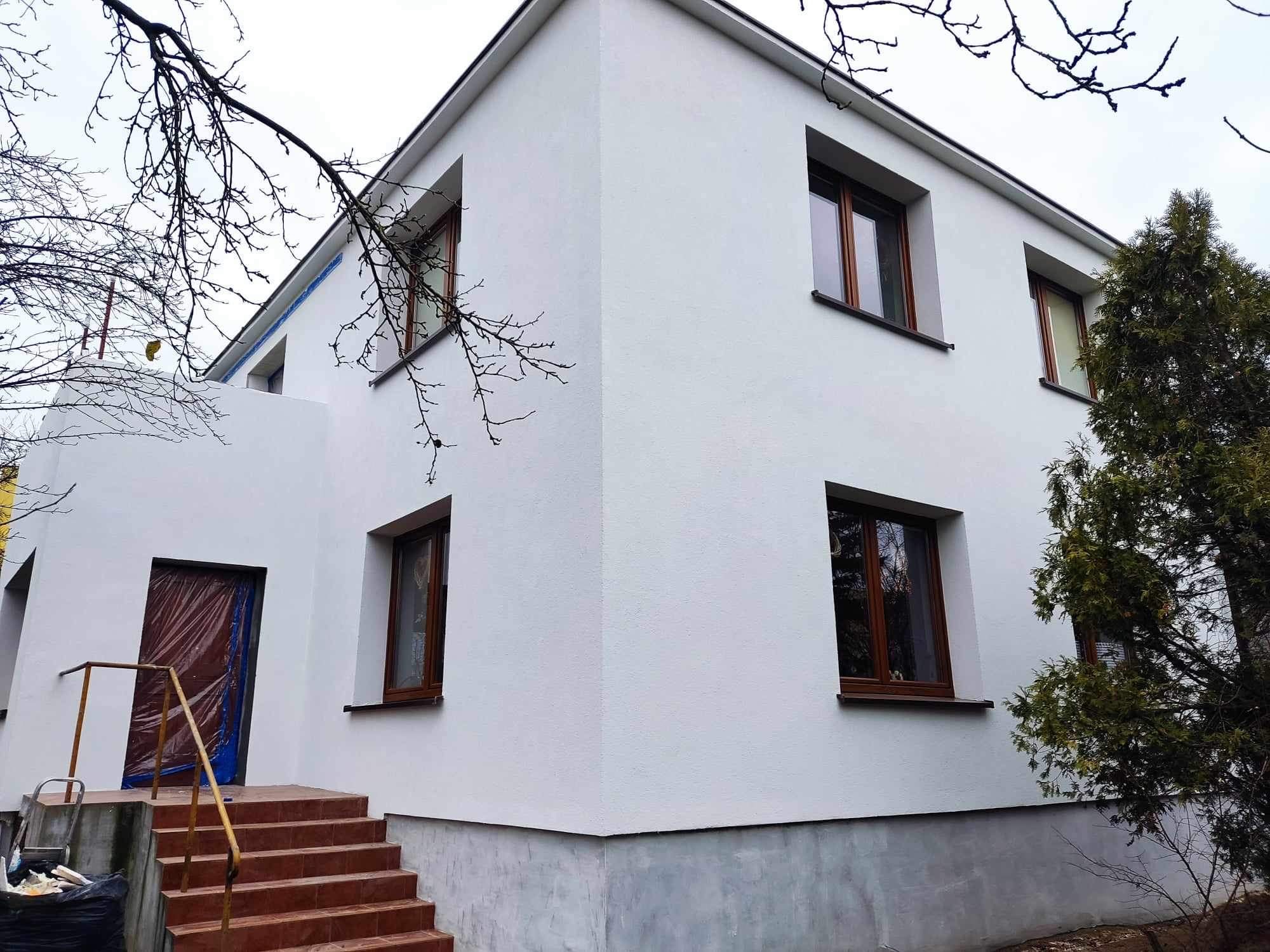 Biała elewacja domu z brązowymi oknami, schody z terakoty prowadzące do drzwi zabezpieczonych folią ochronną, widoczne gałęzie drzew i iglak