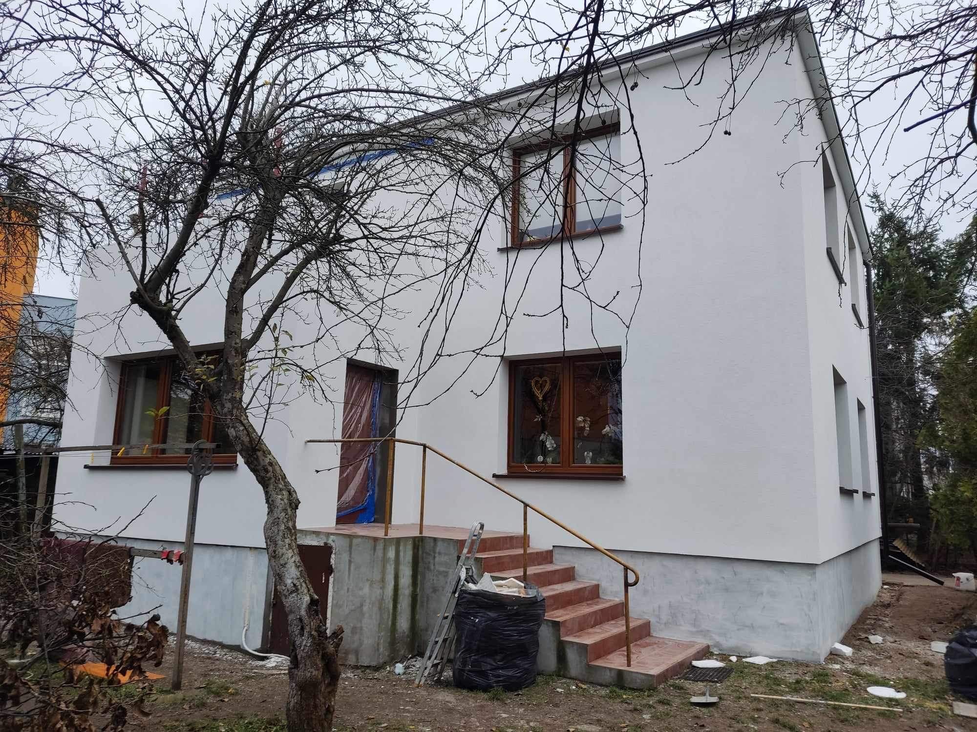 Dwupiętrowy dom z białą elewacją po remoncie, widoczne okna z brązowymi ramami i schody wejściowe z terakoty, na pierwszym planie uschnięte drzewo i śmieci w worku.