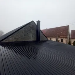 Błyszczący, czarny dach z blachy trapezowej, świeżo po deszczu, z widocznym kominem i fragmentem murowanej ściany szczytowej, na tle zamglonego krajobrazu z budynkami.