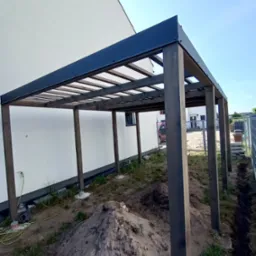 Drewniana pergola z czarnym dachem w trakcie montażu, widoczny nasyp ziemi i przewody elektryczne przy ścianie budynku.