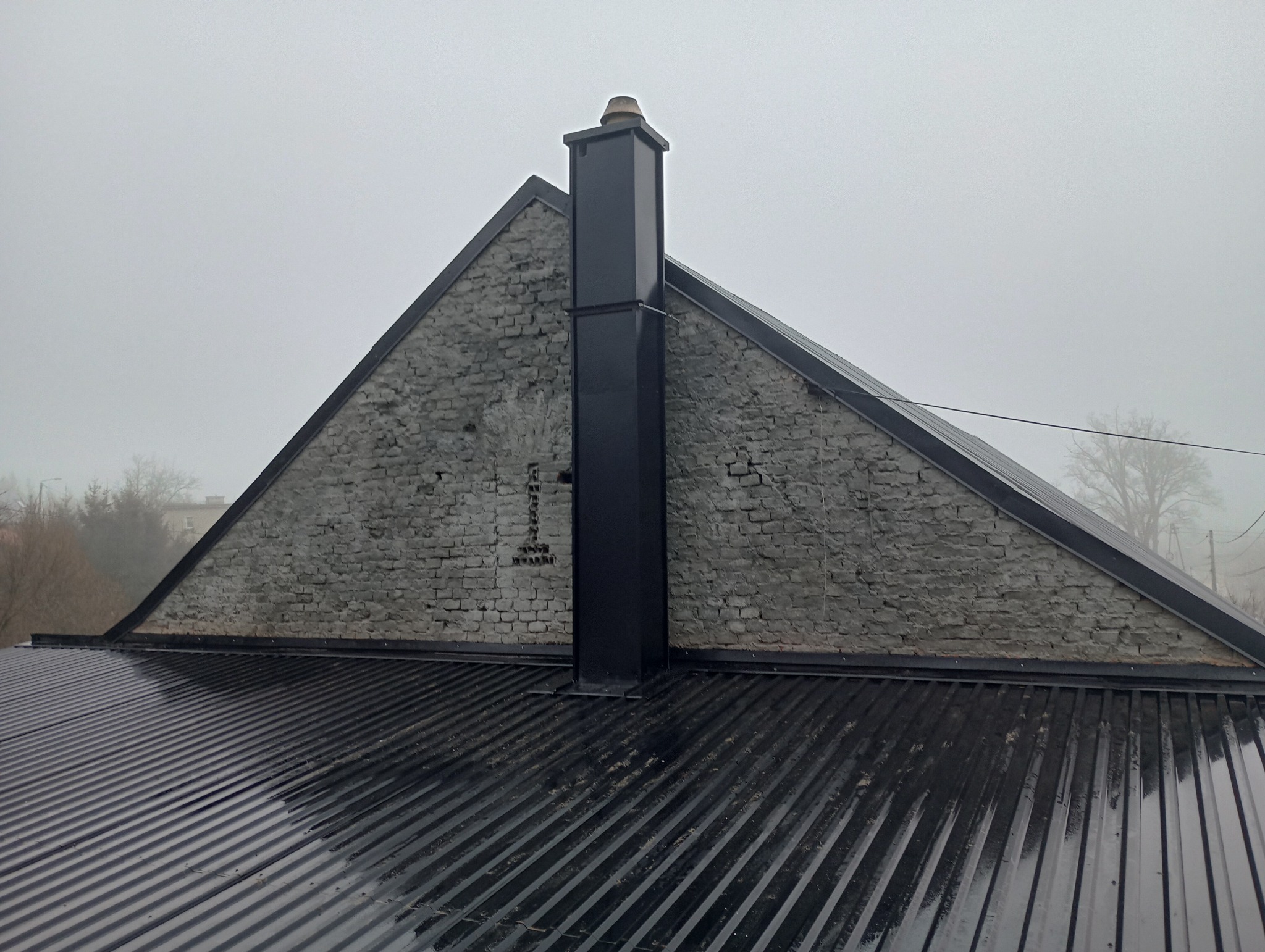 Czarny komin na tle szczytu budynku z cegły, widok z góry na metalowe pokrycie dachu w pochmurny dzień.