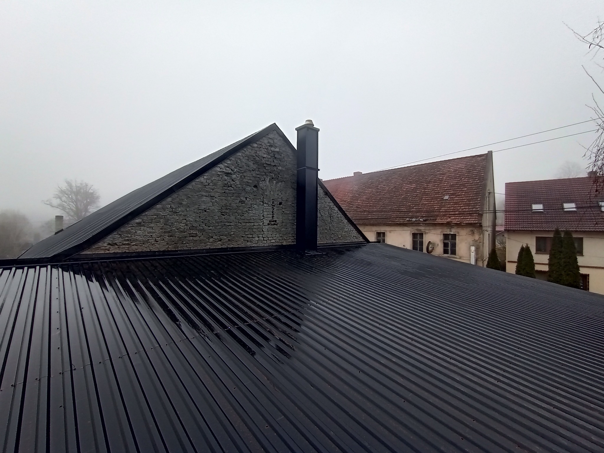 Błyszczący, czarny dach z blachy trapezowej, świeżo po deszczu, z widocznym kominem i fragmentem murowanej ściany szczytowej, na tle zamglonego krajobrazu z budynkami.