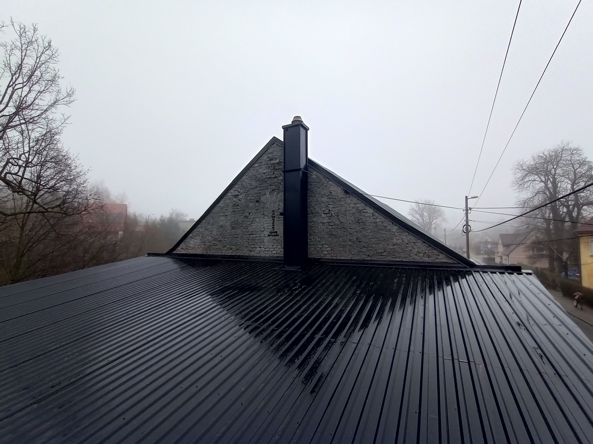 Błyszczący, czarny dach z blachy trapezowej po deszczu, z widocznym kominem na tle mglistego krajobrazu miejskiego i drzew bez liści.