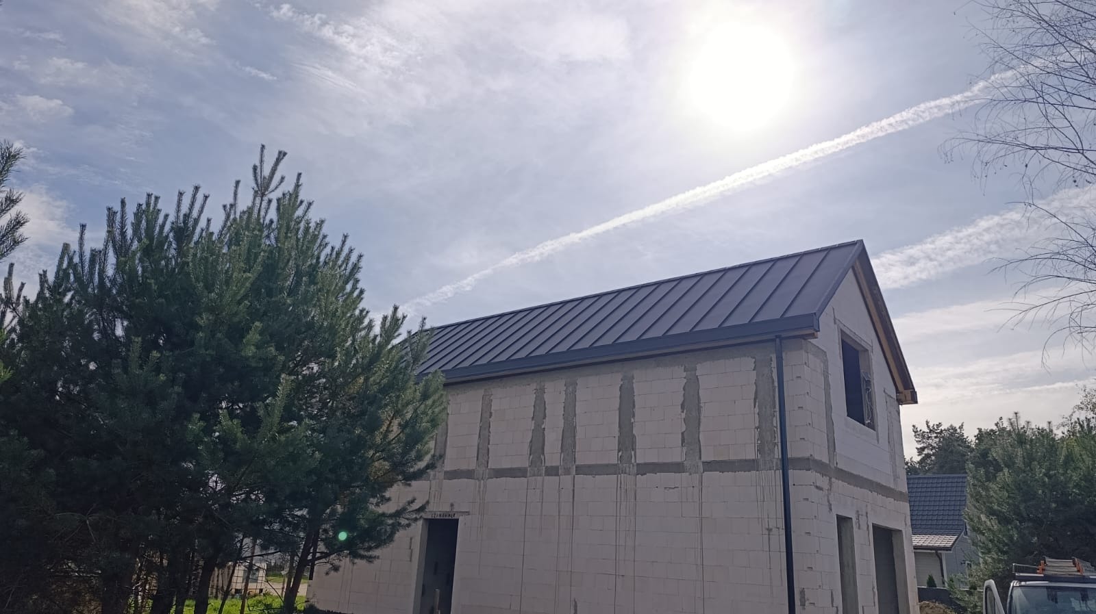 Dom w trakcie budowy z ciemnym, metalowym dachem na tle błękitnego nieba z smugami kondensacyjnymi. Widoczna surowa elewacja i drzewa iglaste.