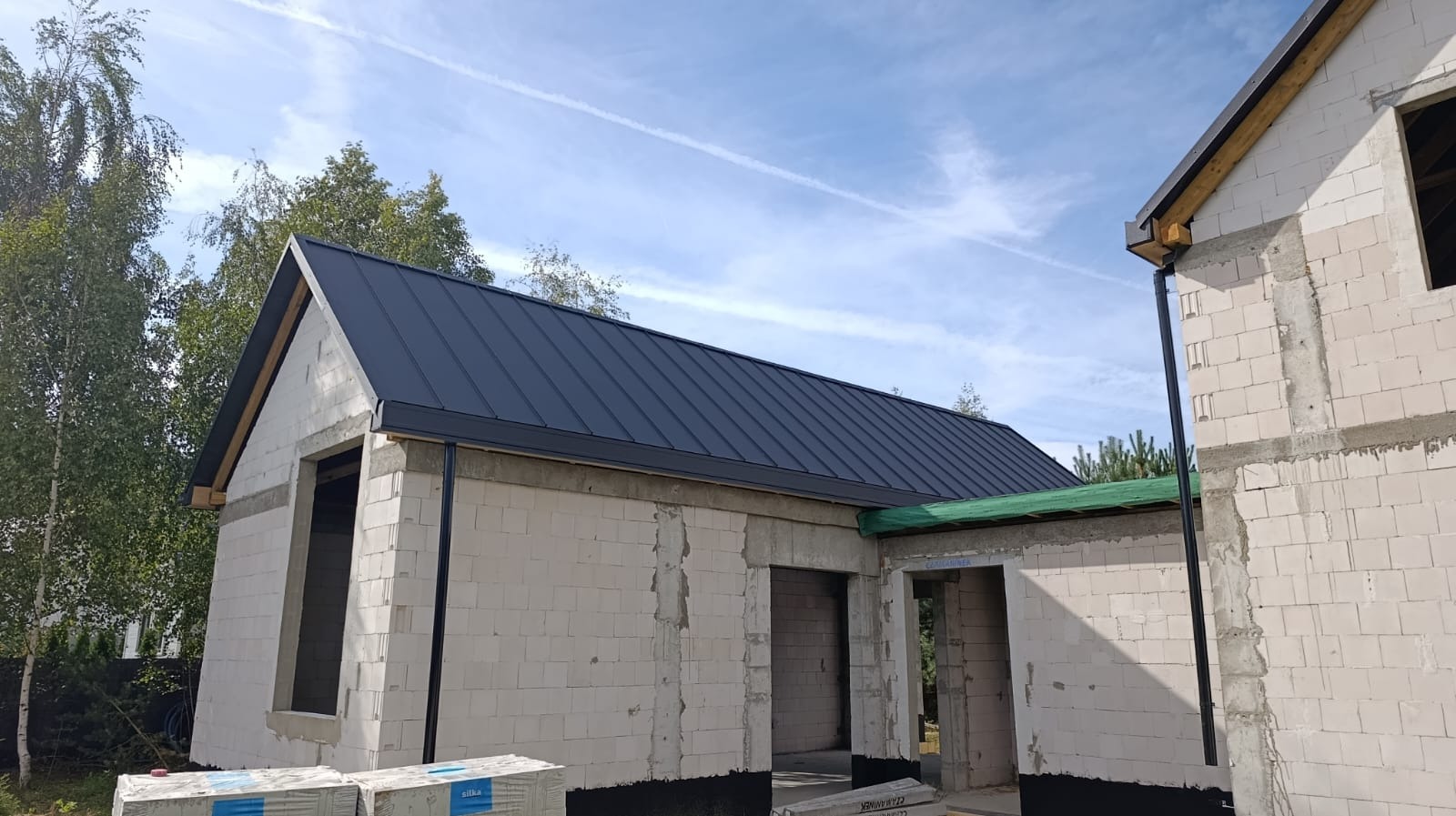 Dom w trakcie budowy z nowym, grafitowym dachem z blachy na rąbek stojący. Widoczne orynnowanie i ściany z betonu komórkowego. W tle drzewa i błękitne niebo.