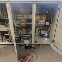 Otwarta szafa sterownicza z widocznymi elementami elektrycznymi, przewodami i miernikiem, na podłodze leżą dodatkowe styczniki.