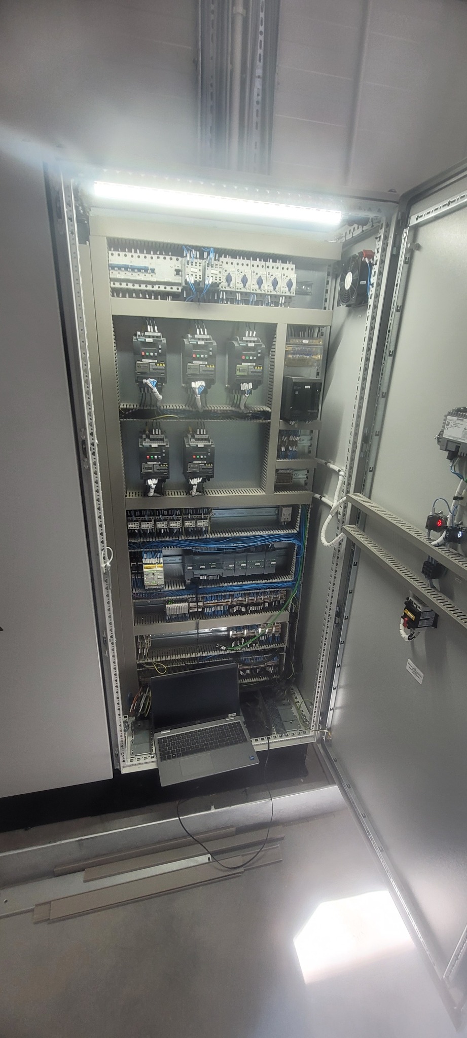 Otwarta szafa sterownicza z zamontowaną aparaturą elektryczną, widoczny laptop podłączony do urządzeń, przewody ułożone w korytkach.