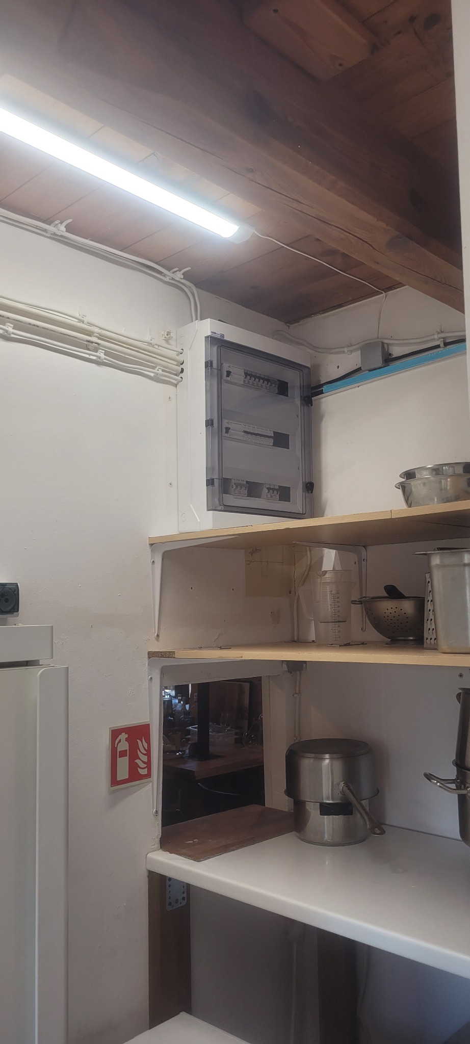 Zamknięta skrzynka z bezpiecznikami, zamontowana na ścianie w pomieszczeniu kuchennym, widoczne kable i rurki instalacyjne, na półkach naczynia kuchenne, drewniany sufit z belkami.