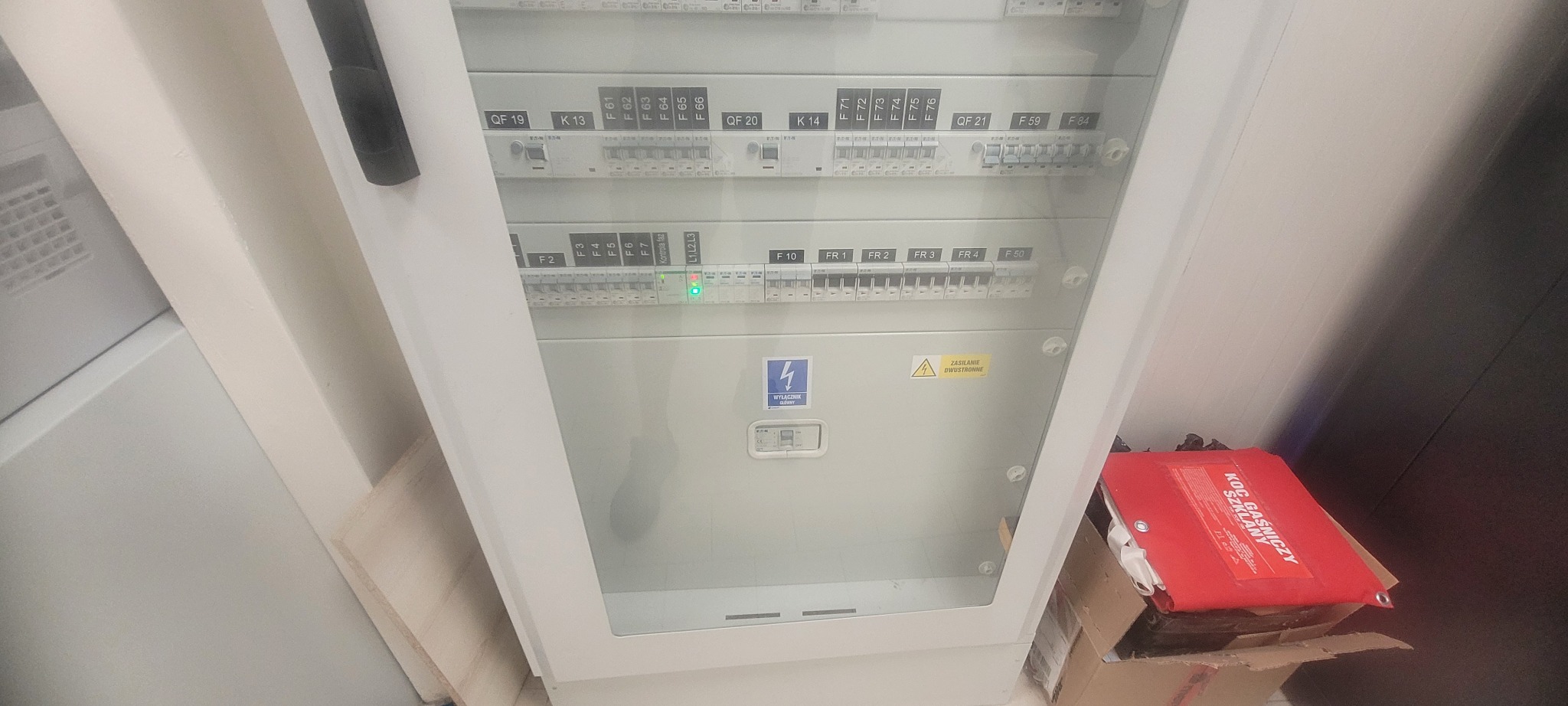Rozdzielnica elektryczna z widocznymi bezpiecznikami i wyłącznikami, z zielonymi diodami sygnalizacyjnymi, obok leży czerwony koc gaśniczy w kartonie.