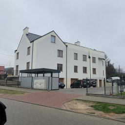 Dardom Dariusz Staniszewski - Nowoczesny budynek wielorodzinny z białymi ścianami i ciemną dachówką, widoczny z ulicy, z zaparkowanymi samochodami i metalowym ogrodzeniem.