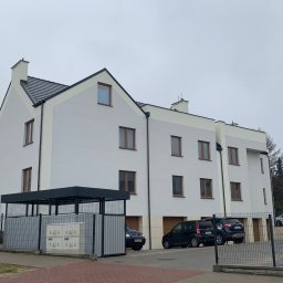 Dardom Dariusz Staniszewski - Nowoczesny budynek wielorodzinny z jasną elewacją i brązowymi oknami, widok z poziomu ulicy, zaparkowane samochody przed budynkiem.