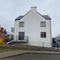 Dardom Dariusz Staniszewski - Dwupiętrowy budynek z białymi ścianami i ciemną dachówką, widoczne brązowe okna i balkony z czarną balustradą, otoczony metalowym ogrodzeniem.