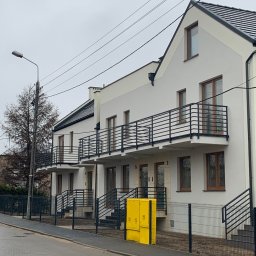 Dardom Dariusz Staniszewski - Nowo wybudowany szeregowiec z białymi elewacjami, czarnymi balustradami balkonowymi i schodami wejściowymi, sfotografowany w pochmurny dzień z perspektywy ulicy, z widoczną żółtą skrzynką gazową...