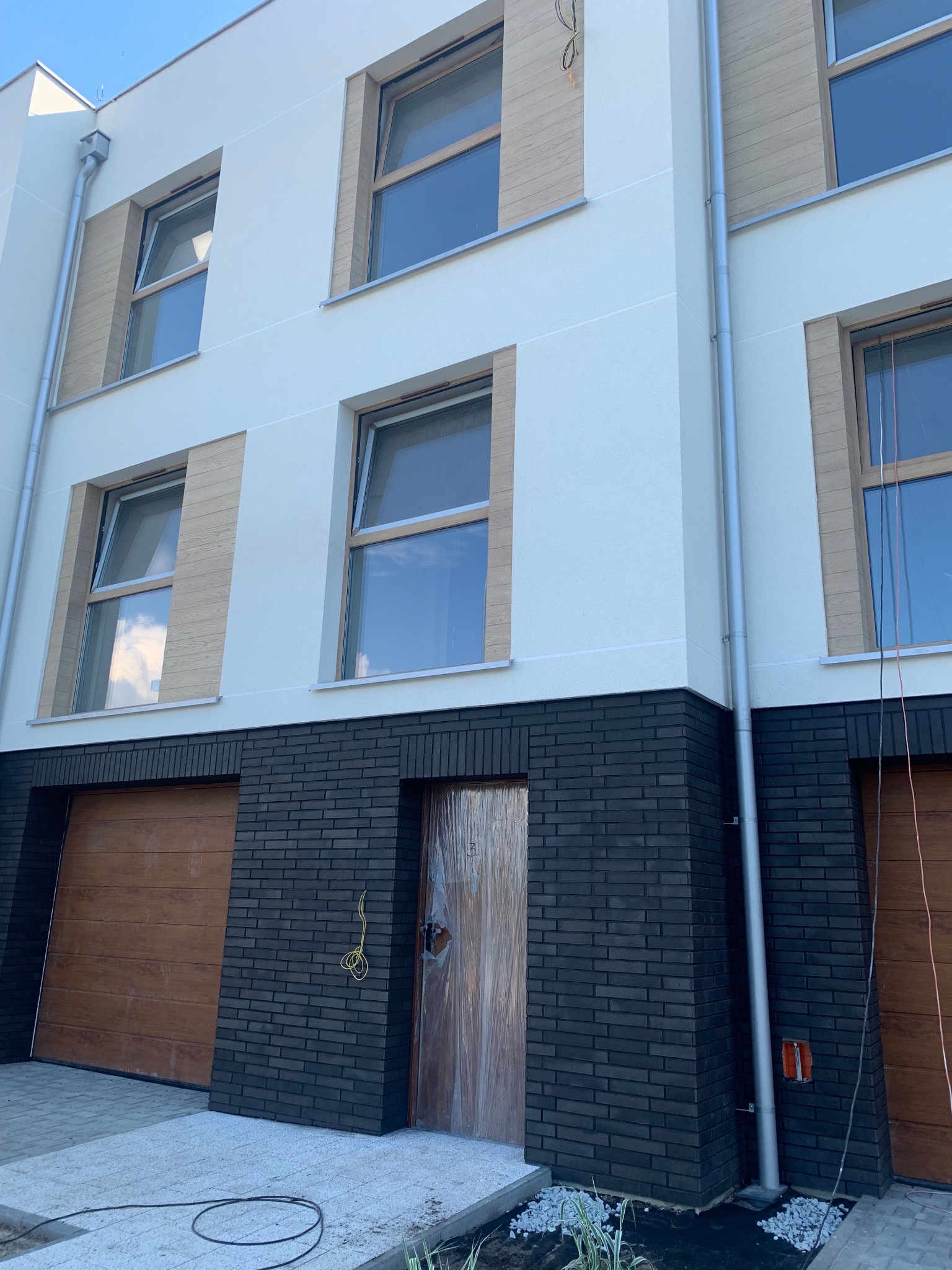 Nowoczesna elewacja budynku z białym tynkiem, drewnopodobnymi panelami okiennymi i ciemną cegłą klinkierową na parterze, widoczne okna i drewniane bramy garażowe.