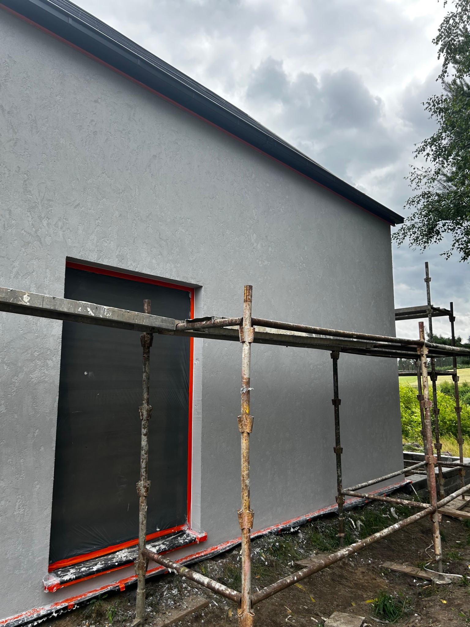 Świeżo otynkowana elewacja budynku w szarym kolorze z widocznym rusztowaniem i zabezpieczonym oknem folią ochronną z pomarańczowym obramowaniem.