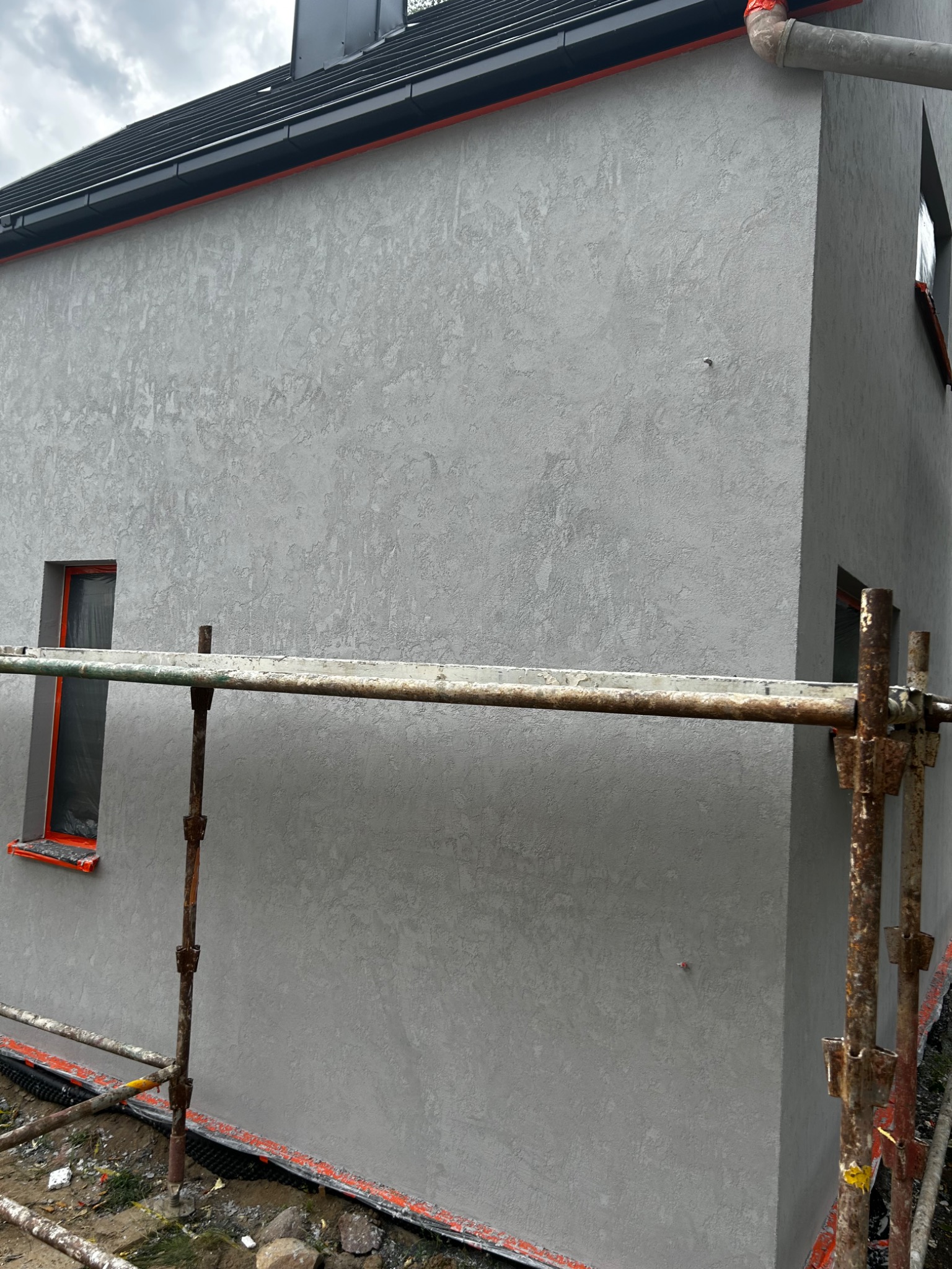 Świeżo otynkowana szara elewacja domu jednorodzinnego z widocznymi rusztowaniami i zabezpieczonymi oknami folią malarską.