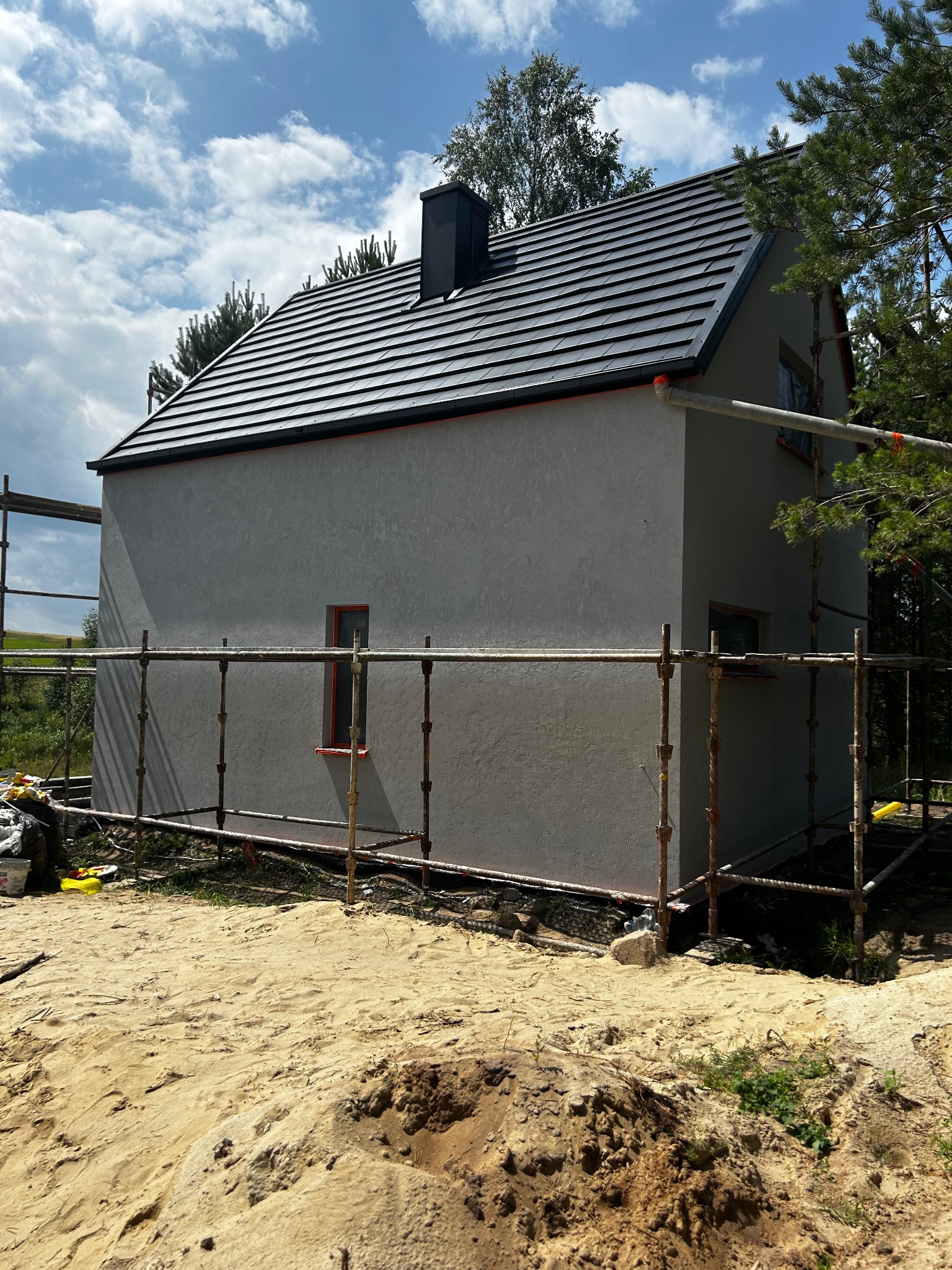 Dom jednorodzinny w trakcie ocieplania, elewacja pokryta szarą strukturą, rusztowania wzdłuż ściany, czarny dach z dachówki ceramicznej, okna z czerwonymi ramami.