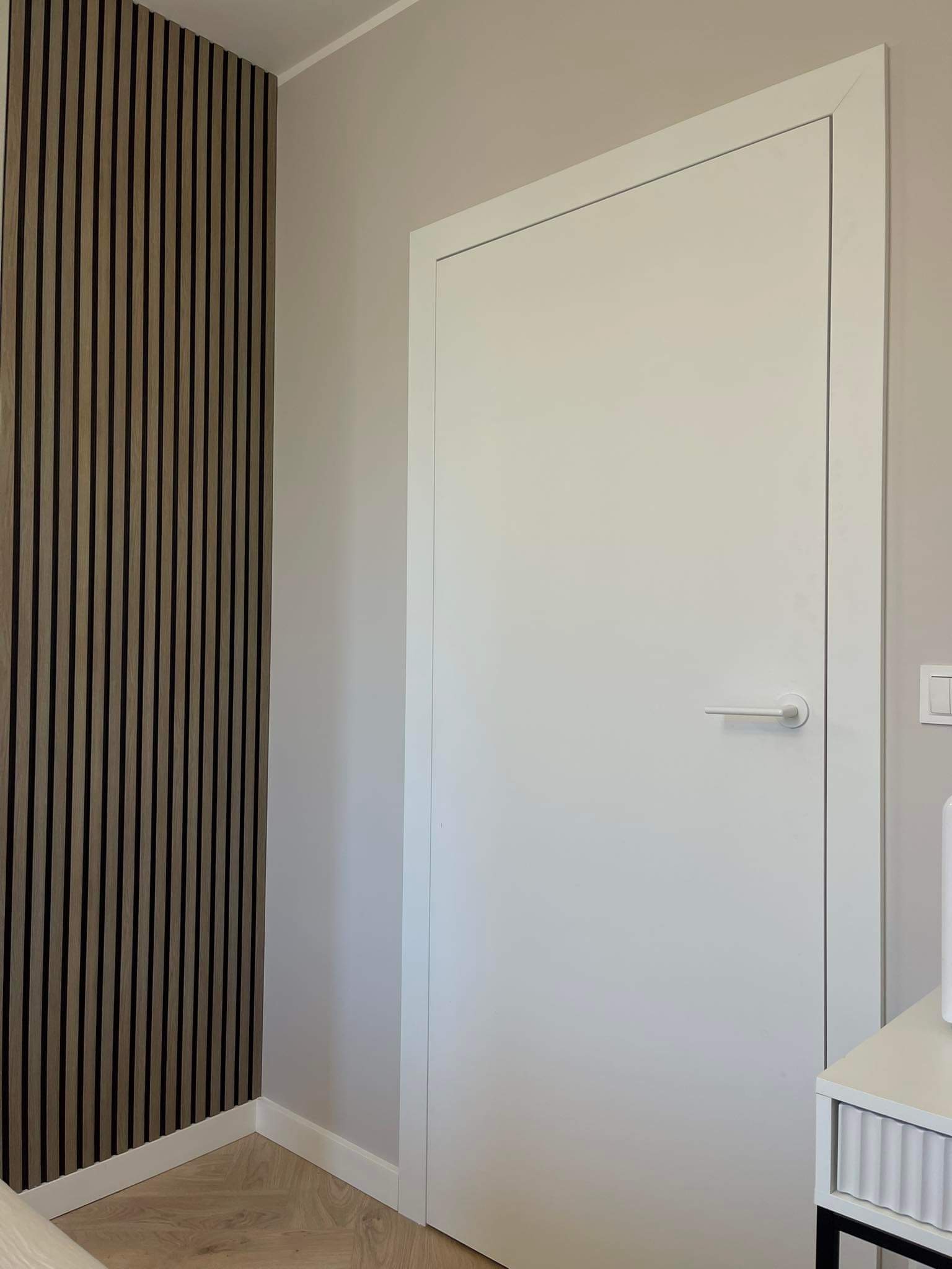 Nowoczesne wnętrze: białe drzwi z minimalistyczną klamką na tle ściany z drewnianych paneli akustycznych w odcieniach beżu i brązu, widoczny fragment parkietu w jodełkę.
