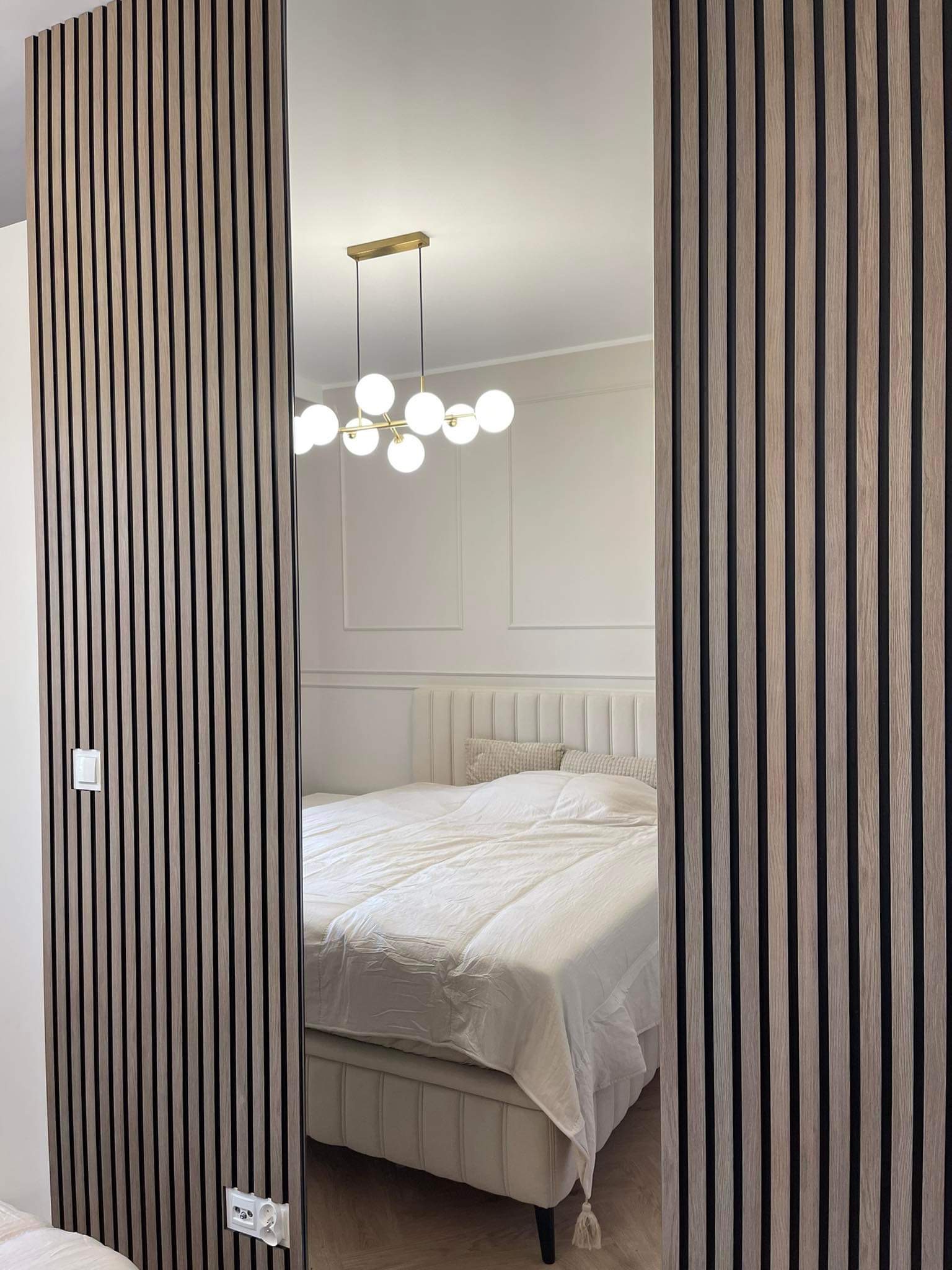 Drewniane lamele ścienne w sypialni z widocznym gniazdkiem elektrycznym i włącznikiem światła, dekoracyjne listwy na ścianie i sufitowa lampa z kulistymi kloszami.