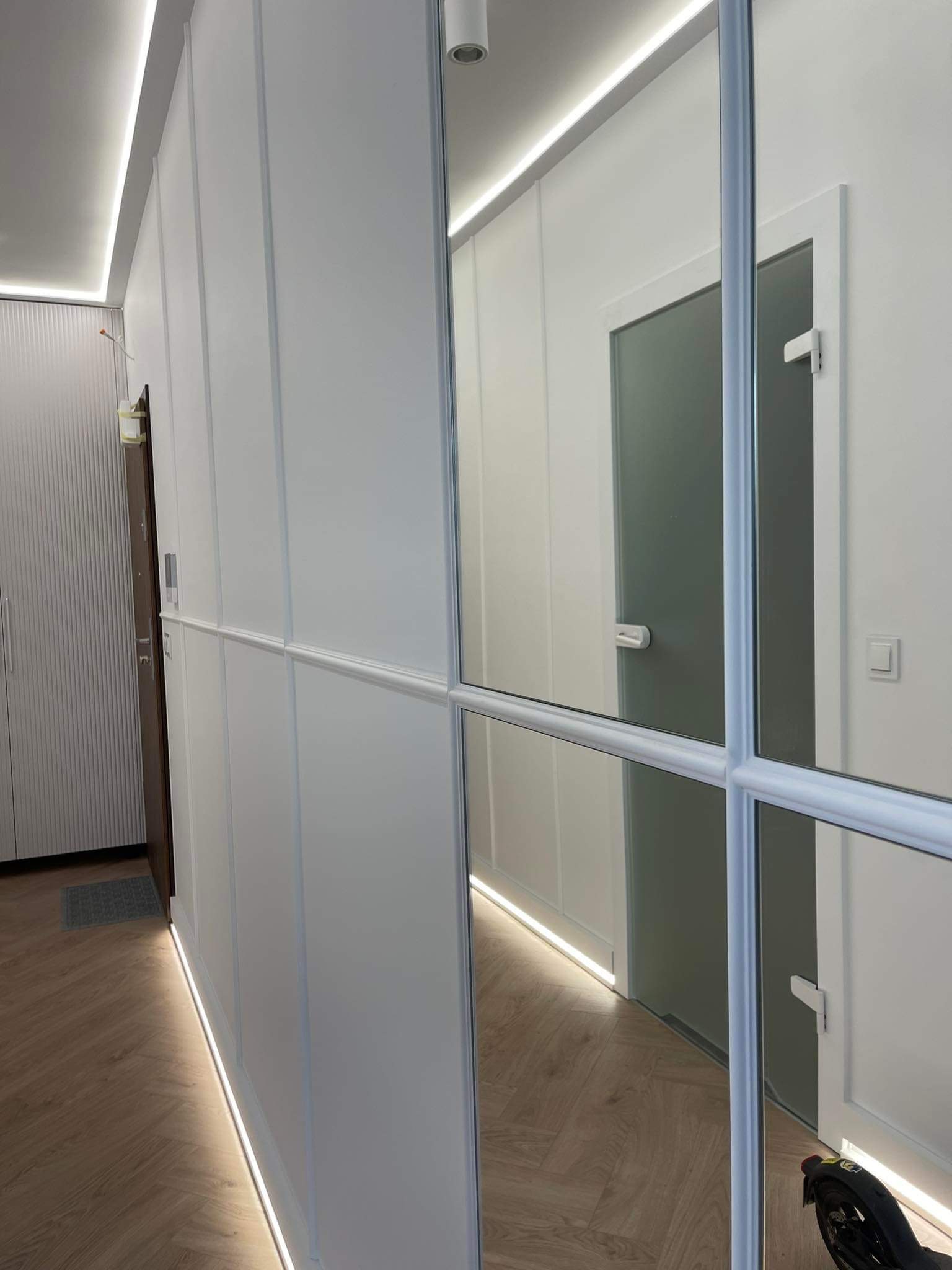 Nowoczesny przedpokój z białymi panelami ściennymi z subtelnym oświetleniem LED przy podłodze i suficie, lustrzane drzwi z metalową ramą, parkiet w jodełkę.