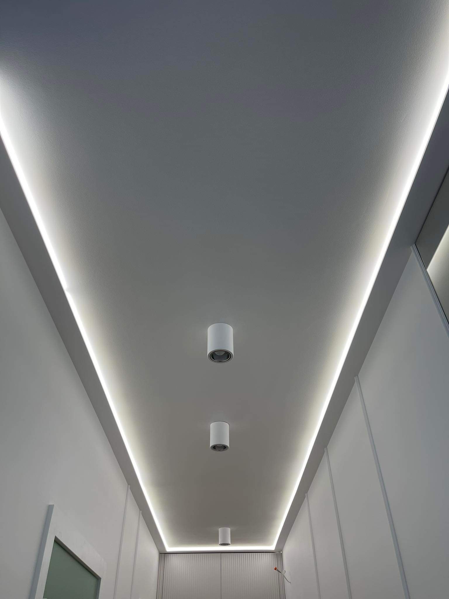 Biały sufit z wbudowanym oświetleniem LED wzdłuż krawędzi i trzema punktowymi lampami sufitowymi, minimalistyczny design.