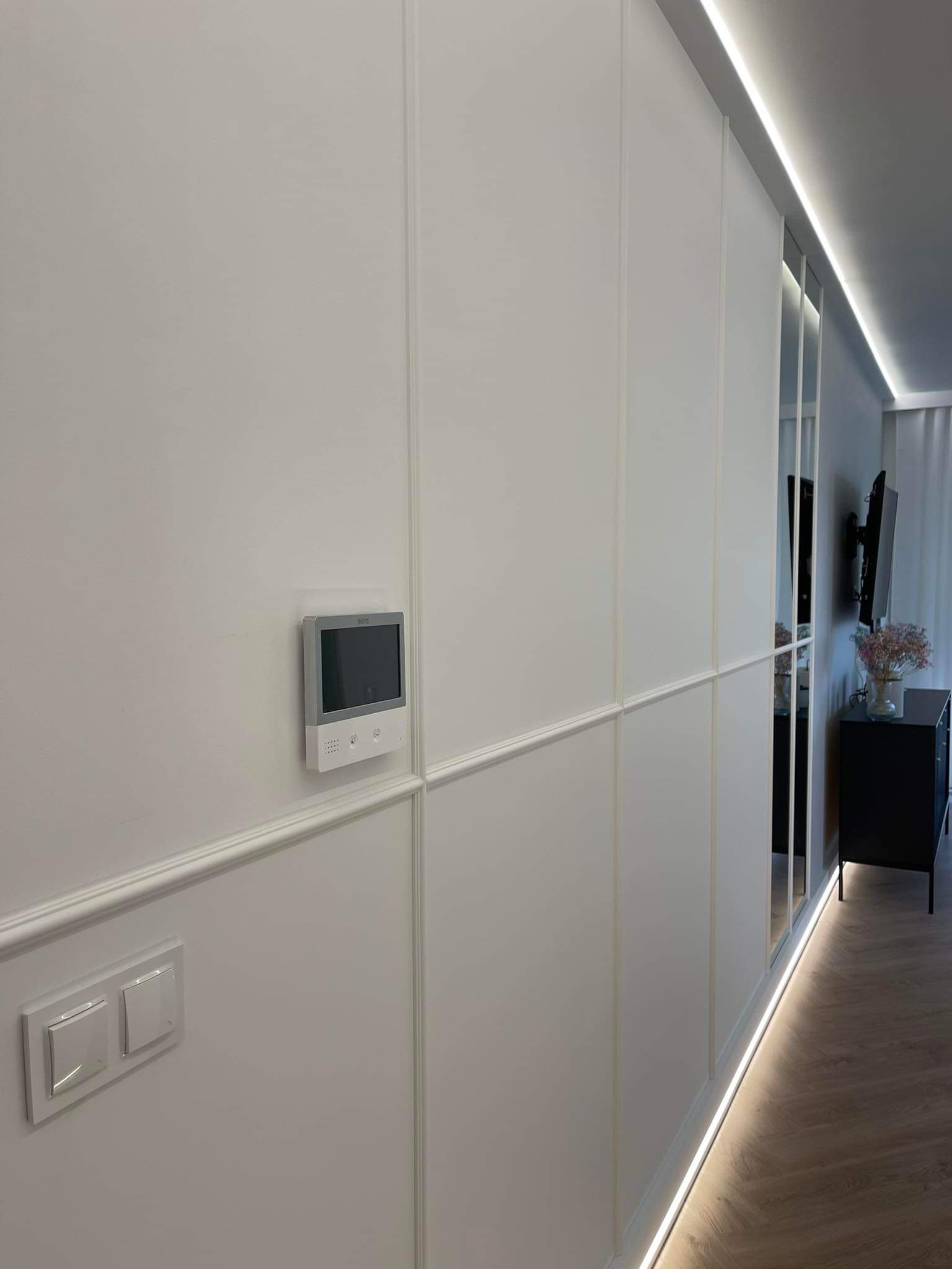 Biała ściana z panelami dekoracyjnymi, wbudowanym oświetleniem LED przy podłodze i suficie, włącznik światła i domofon, widok na fragment wnętrza z szafką i telewizorem.