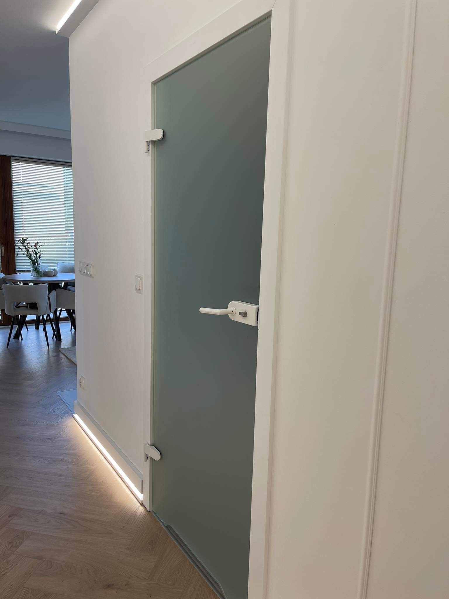 Szklane drzwi wewnętrzne z białymi ościeżnicami i klamką, oświetlenie LED przy podłodze, parkiet w jodełkę, widok na jadalnię z żaluzjami.