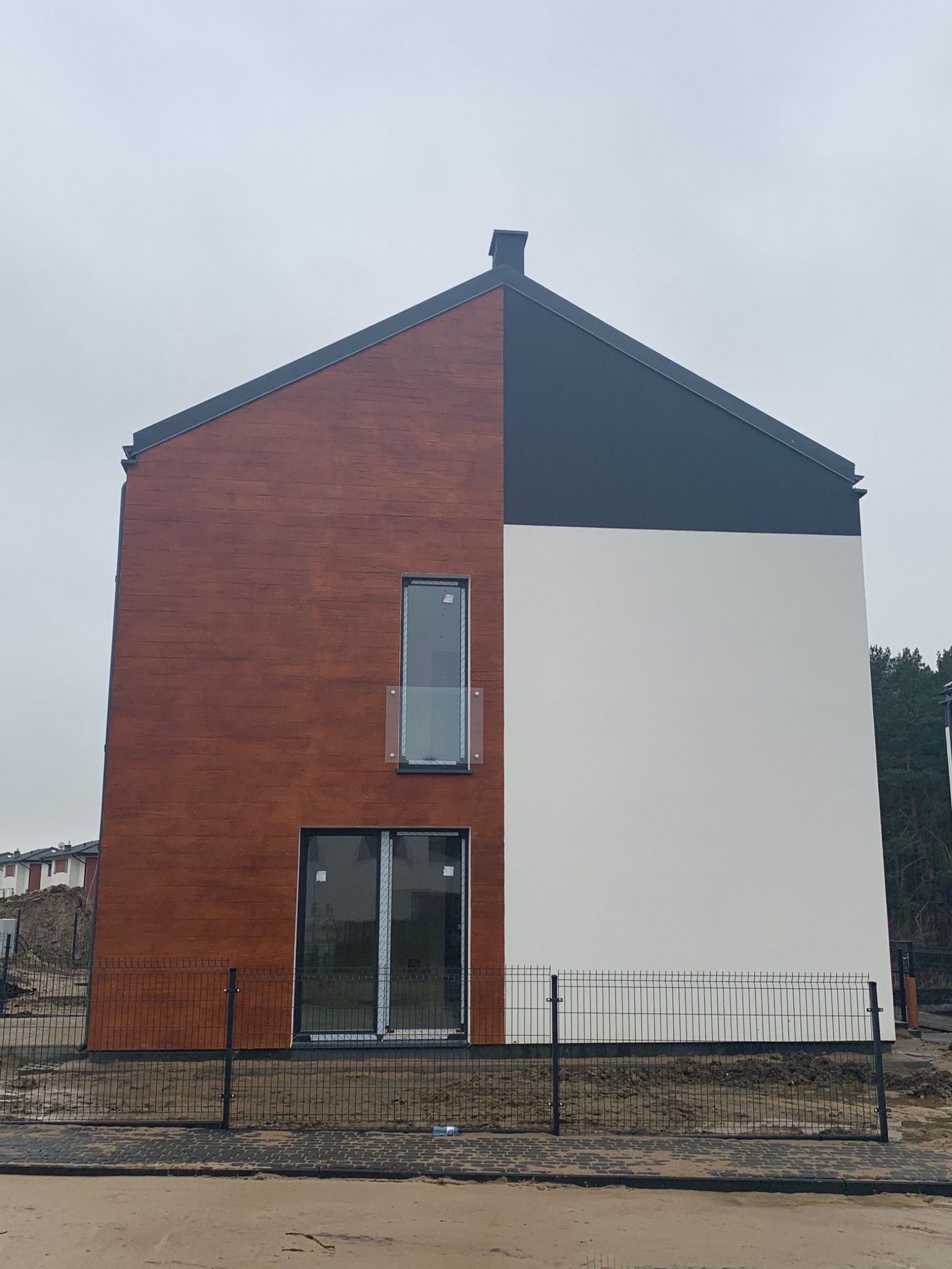 Nowoczesny dom jednorodzinny z elewacją w kolorze drewna i białym tynkiem, czarny dach, widok z zewnątrz na ogrodzenie i teren w trakcie zagospodarowania.