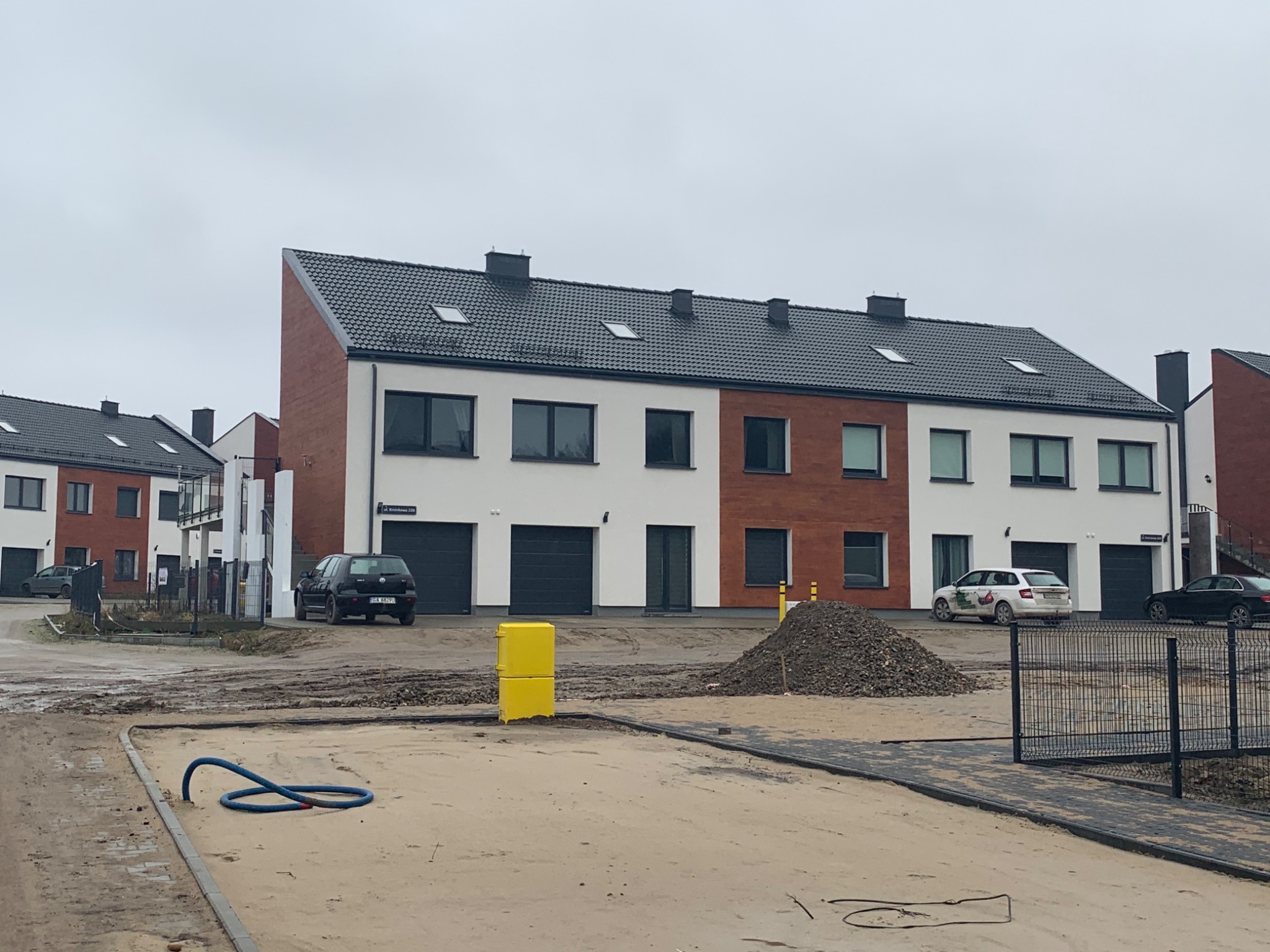 Nowo wybudowane szeregówki z białymi i ceglanymi elewacjami, widoczne samochody, żółta skrzynka i hałda ziemi na placu budowy.