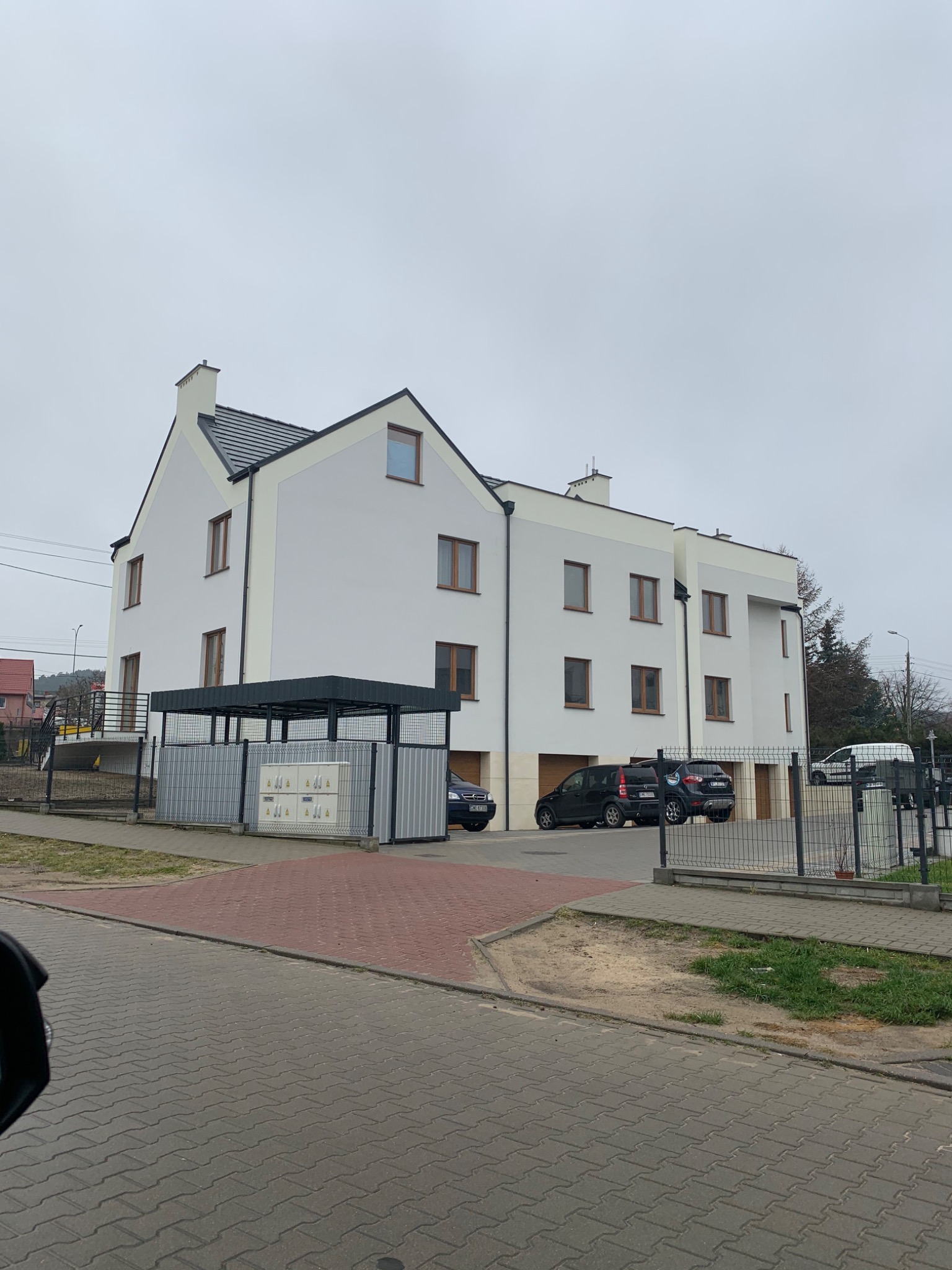Nowoczesny budynek wielorodzinny z białymi ścianami i ciemną dachówką, widoczny z ulicy, z zaparkowanymi samochodami i metalowym ogrodzeniem.