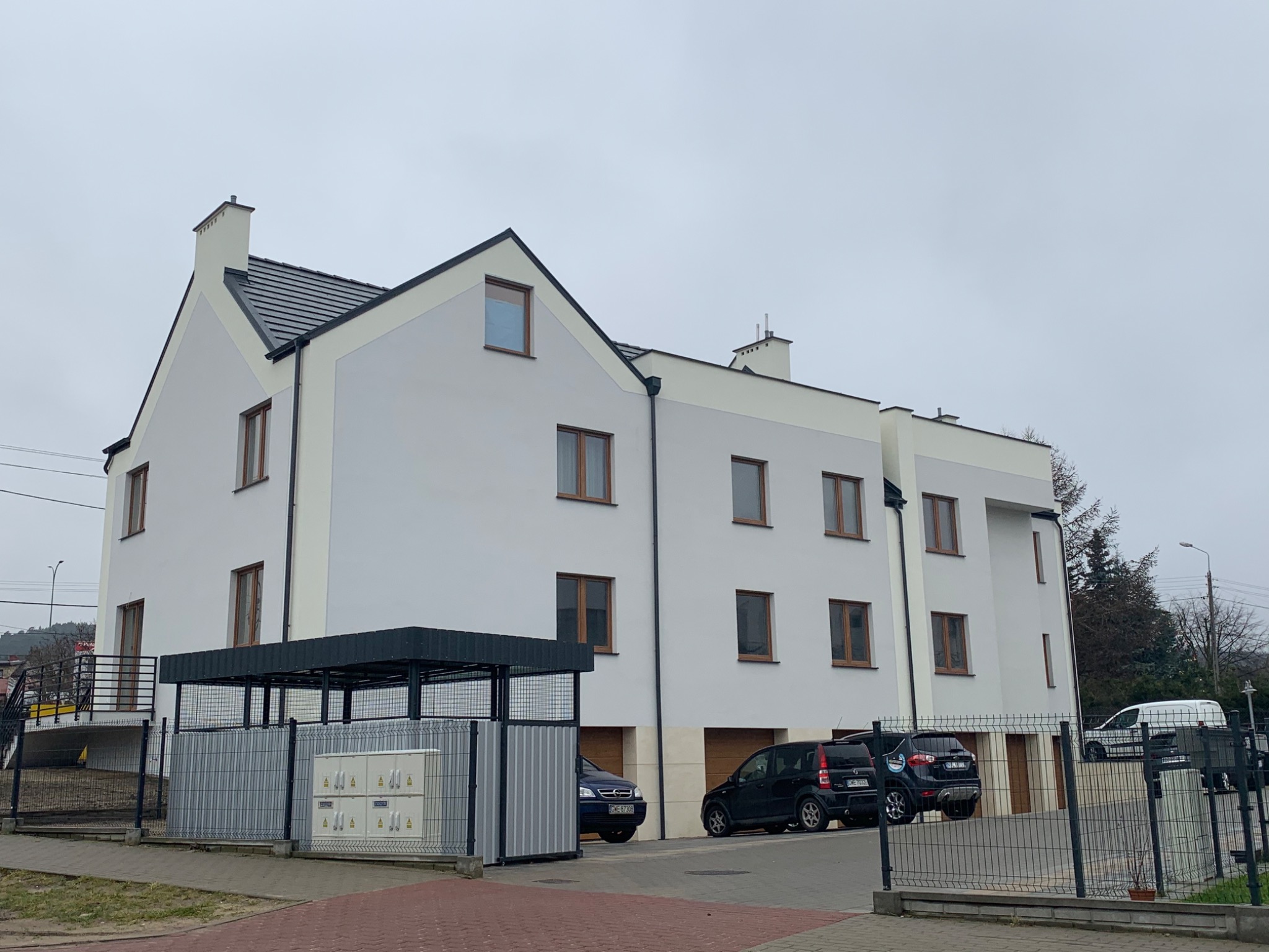 Nowoczesny budynek wielorodzinny z jasną elewacją i brązowymi oknami, widok z poziomu ulicy, zaparkowane samochody przed budynkiem.