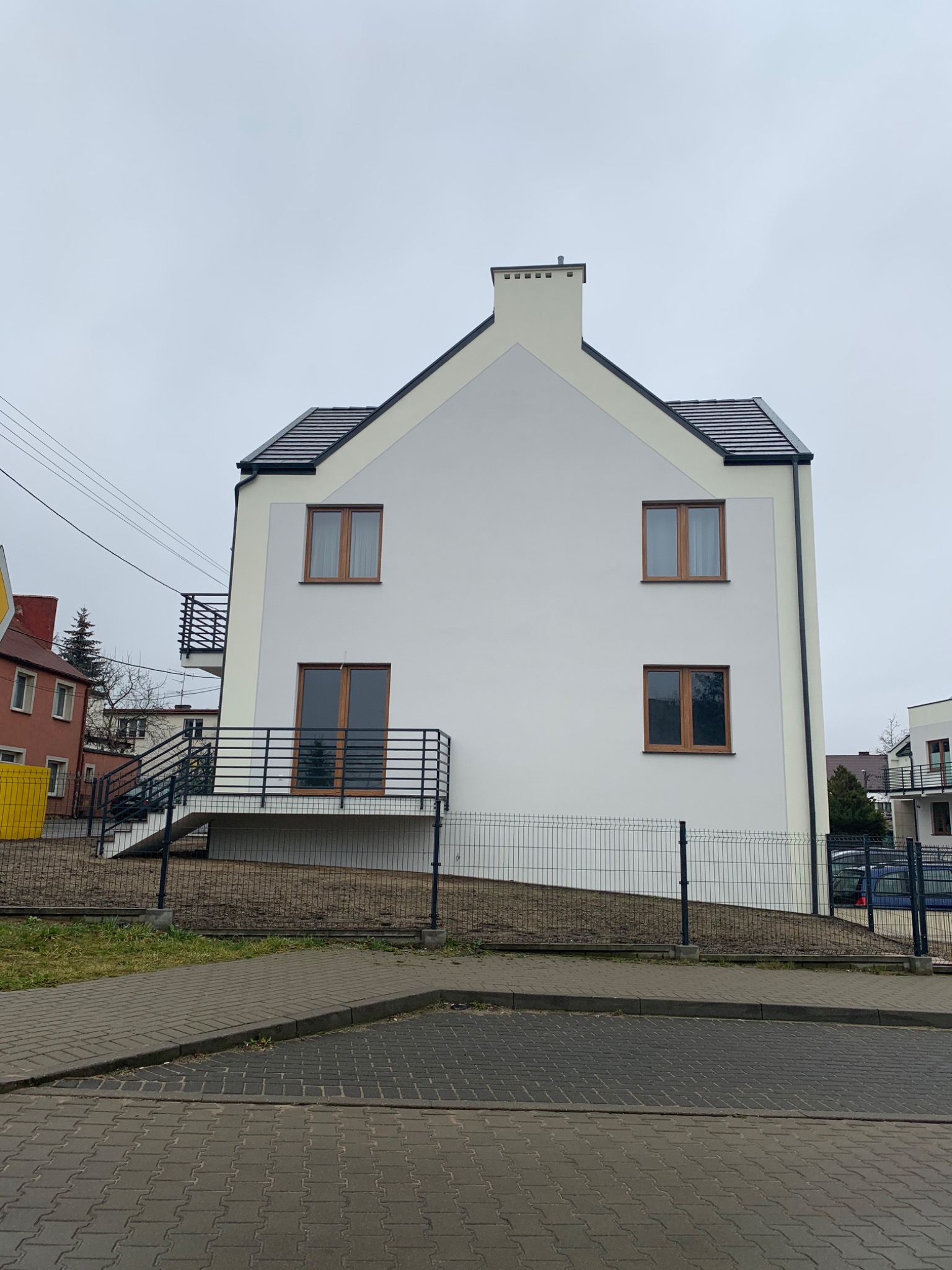 Dwupiętrowy budynek z białymi ścianami i ciemną dachówką, widoczne brązowe okna i balkony z czarną balustradą, otoczony metalowym ogrodzeniem.