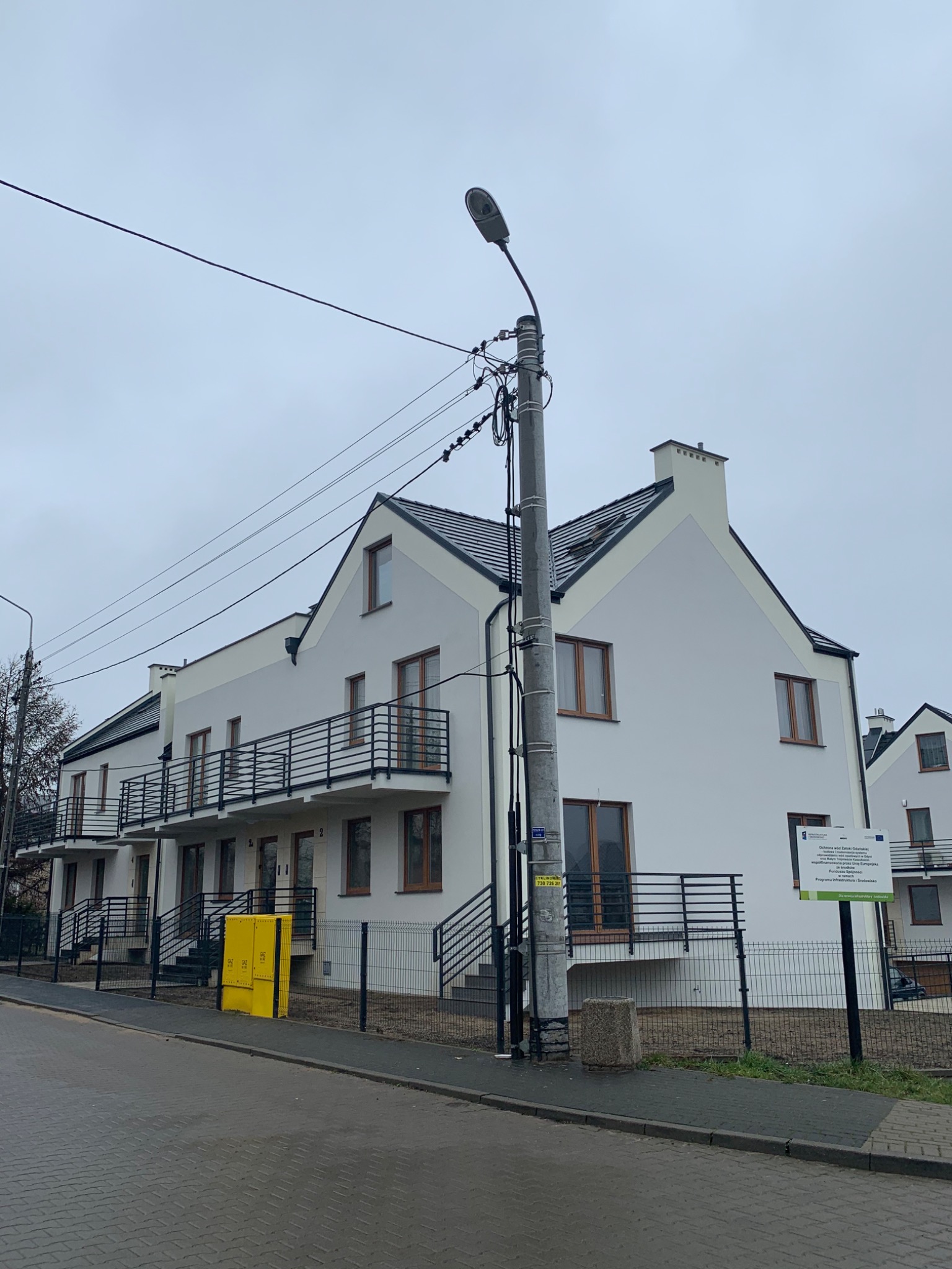 Nowe, pomalowane na biało budynki wielorodzinne z balkonami i brązowymi oknami, widoczne z poziomu ulicy w pochmurny dzień, z żółtą skrzynką gazową i słupem elektrycznym na pierwszym planie.